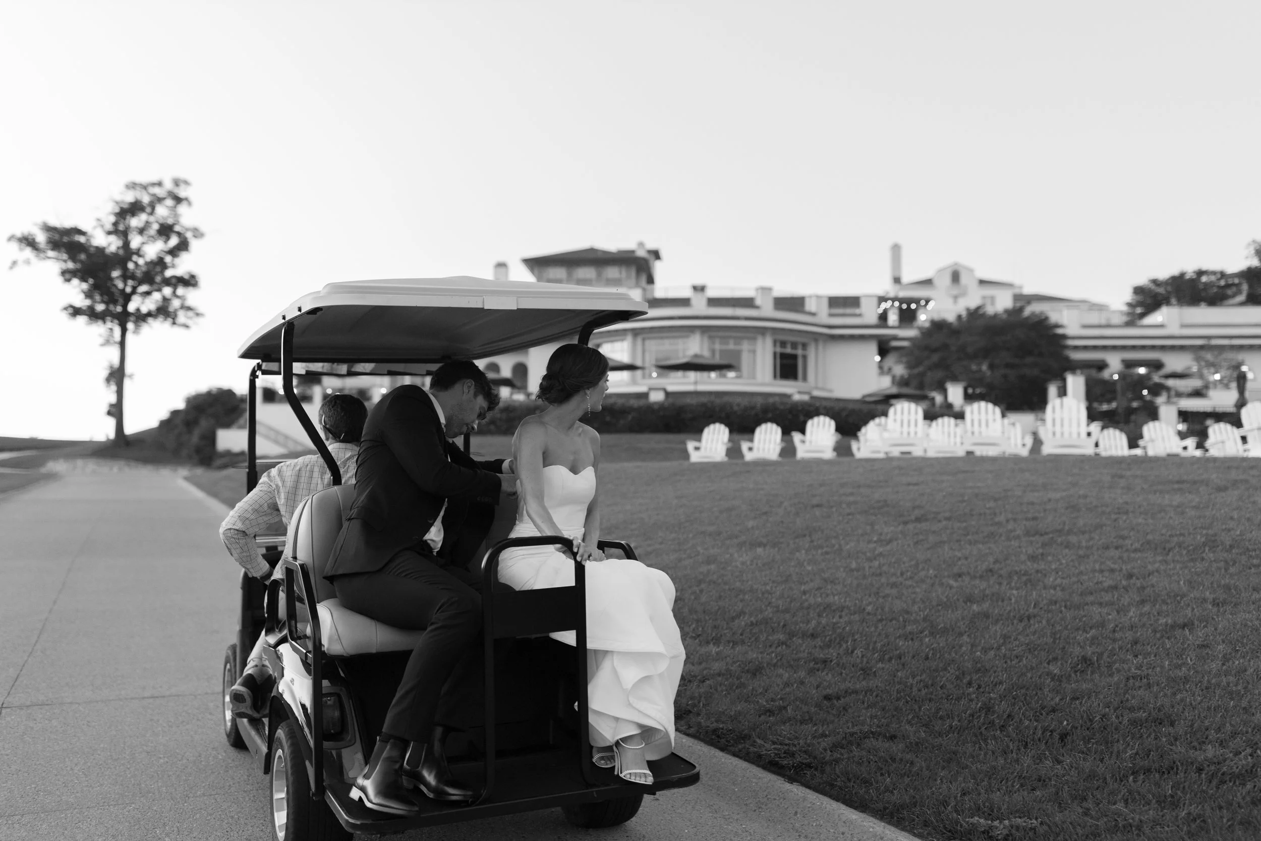 A bride in a white wedding dress sits in a golf cart with three men, one in a tuxedo. The golf cart is driving on a paved path near a large house and lawn with Adirondack chairs in the background.