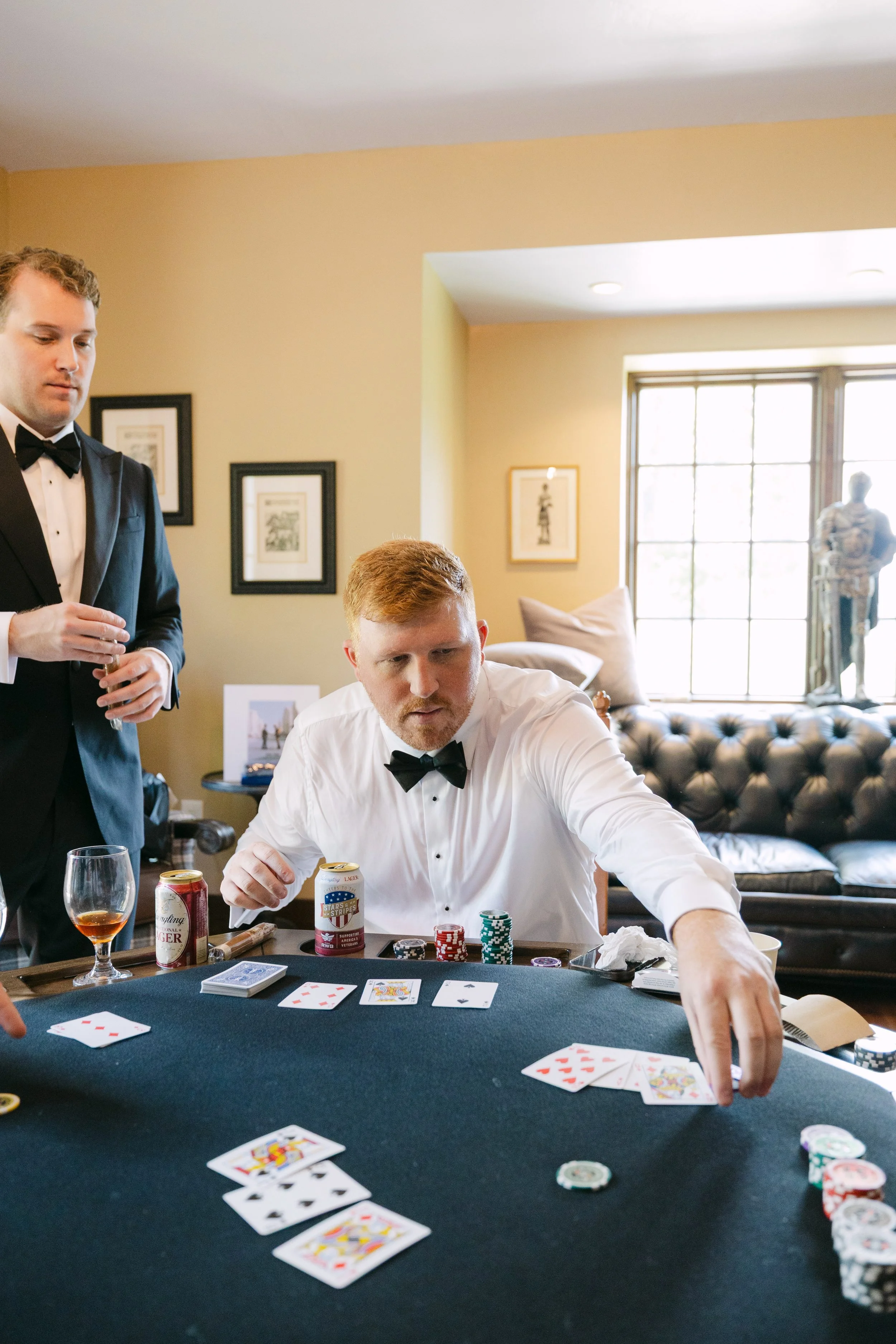 Two men in tuxedos playing poker at a game table in a well-lit living room with artwork on the walls and a large window.