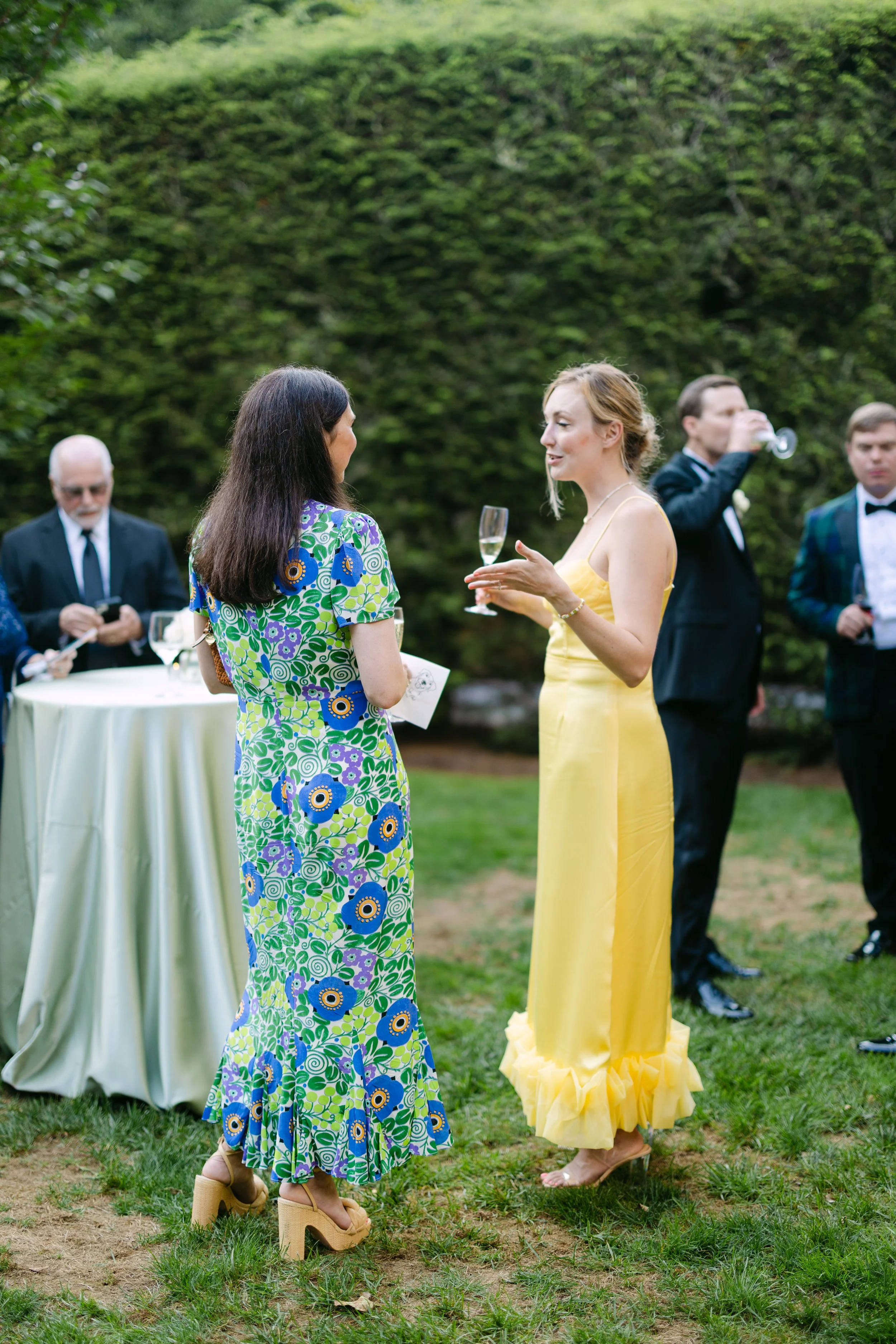 Two women are talking and drinking at an outdoor event, with other people in formal attire in the background.