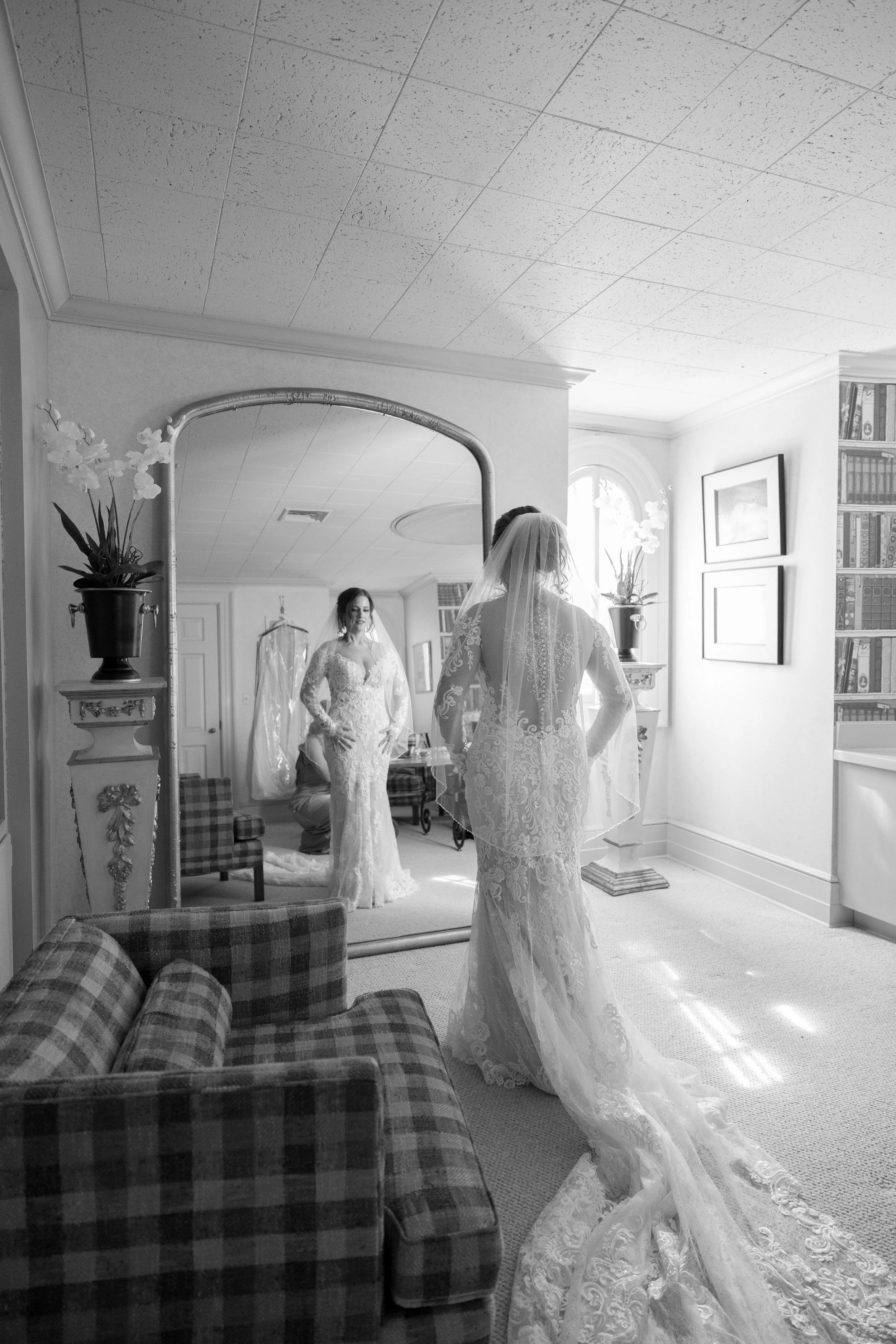 A bride in a lace wedding dress and veil stands before a large mirror in a bright, elegantly decorated room, preparing for her wedding.