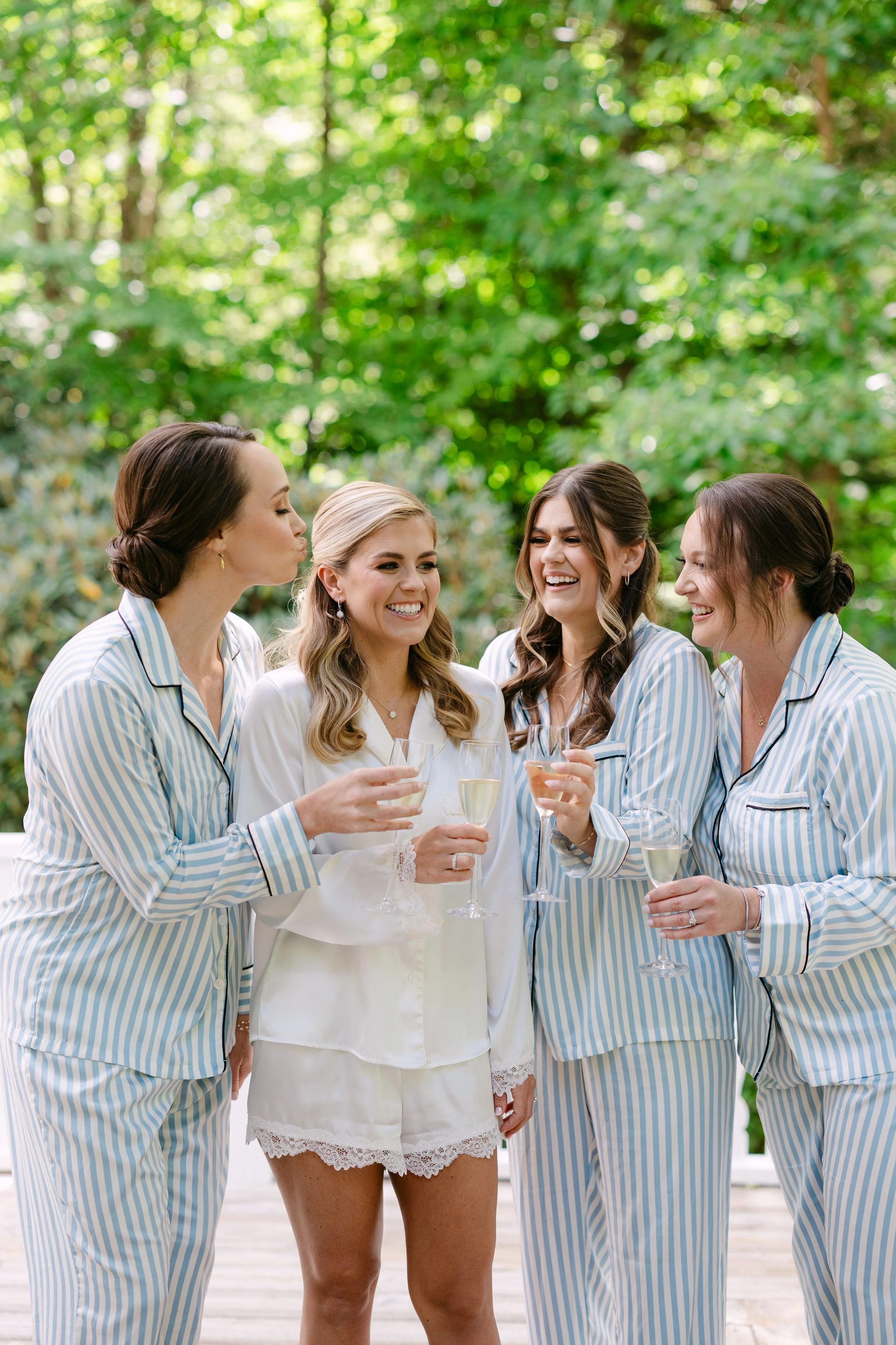 Four women in pajama sets holding glasses of champagne and toasting outdoors in a lush, green forest.