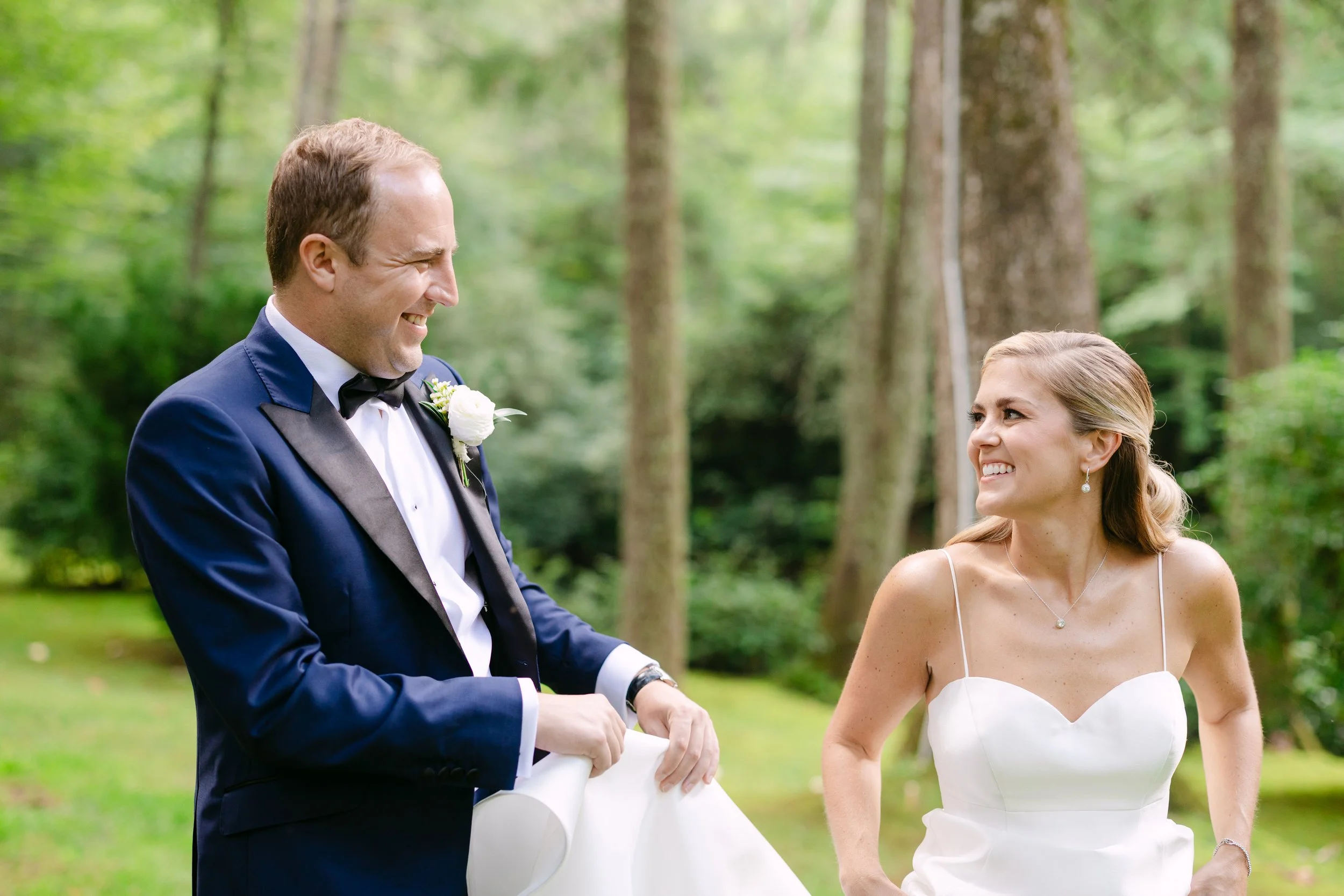 A newlywed couple standing outdoors in a wooded area, smiling at each other. The groom is dressed in a navy tuxedo with a white shirt and black bow tie, holding a white fabric. The bride is wearing a white strapless wedding dress with thin spaghetti 