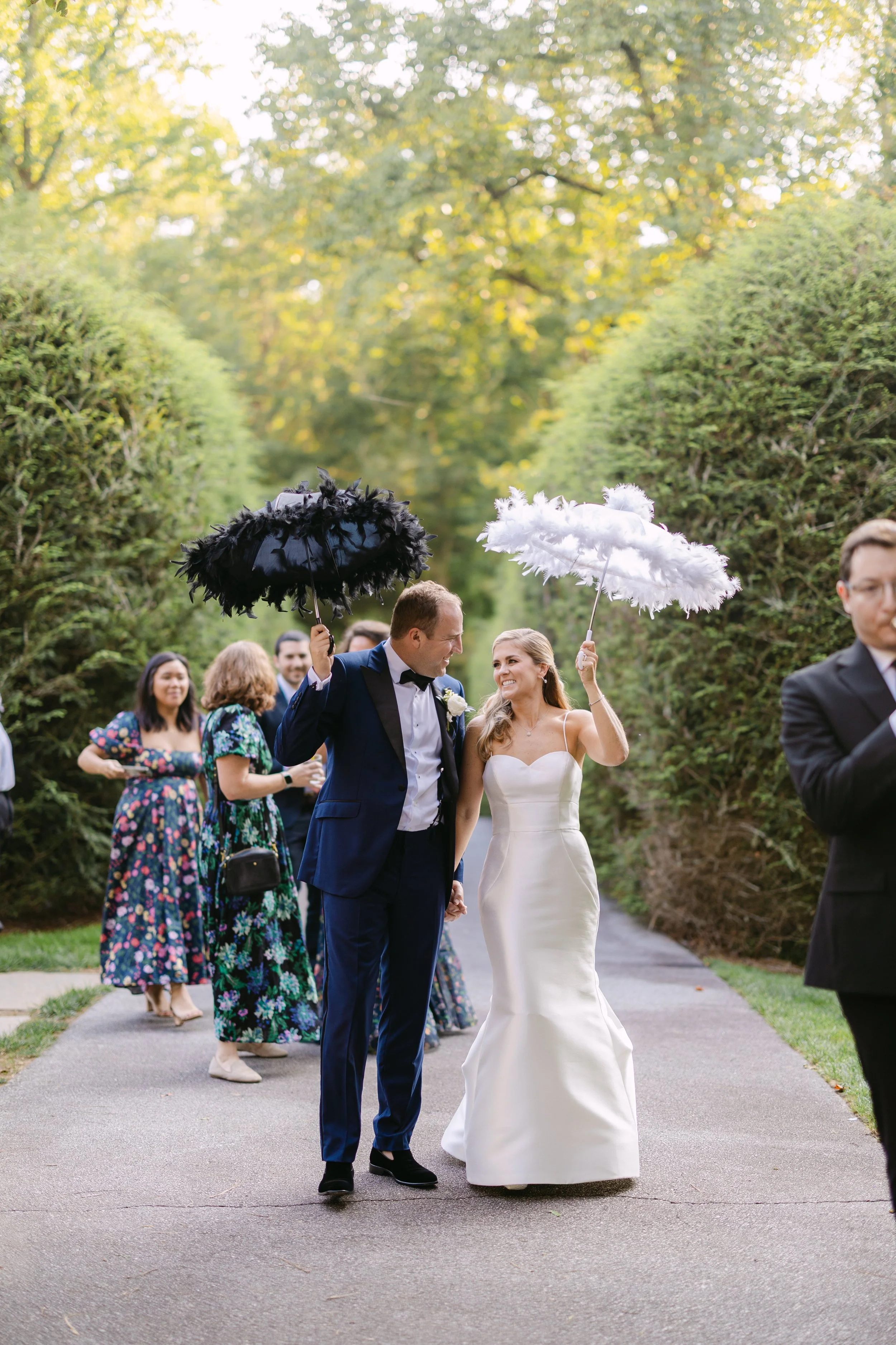 A bride and groom holding hands while walking outside, smiling at each other, with guests in the background holding black and white feather umbrellas.