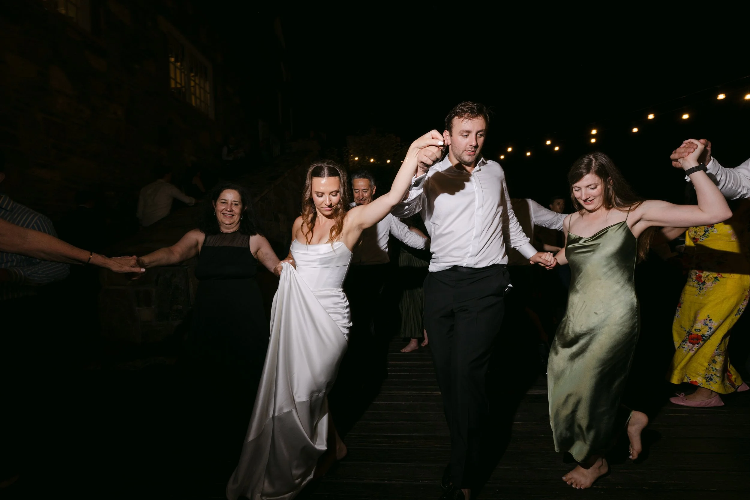People dancing in a circle at a celebration or wedding during nighttime, holding hands and smiling.