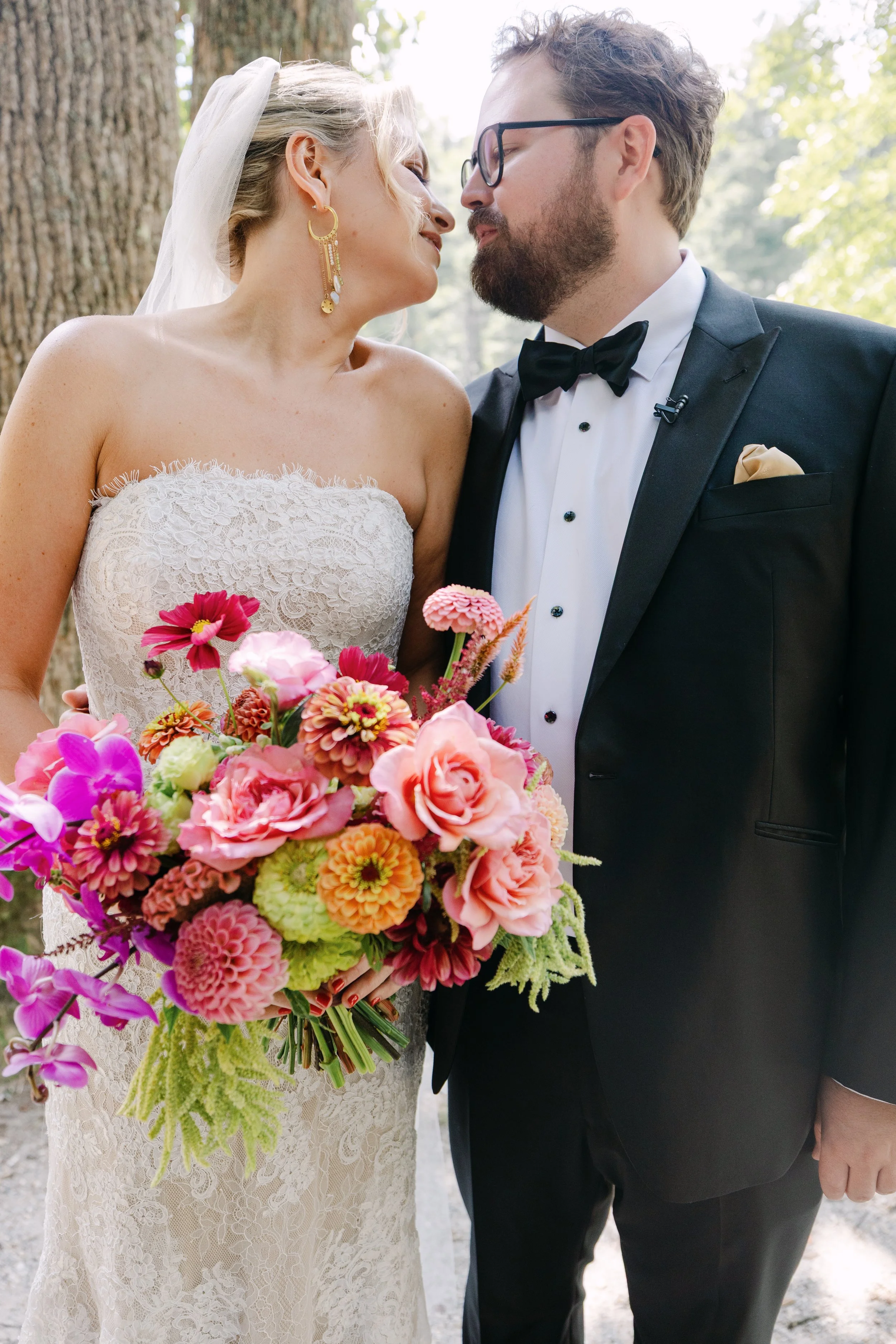 Bride and groom in wedding attire, about to kiss, outdoors with large trees, the bride holding a vibrant bouquet of pink, peach, and purple flowers.