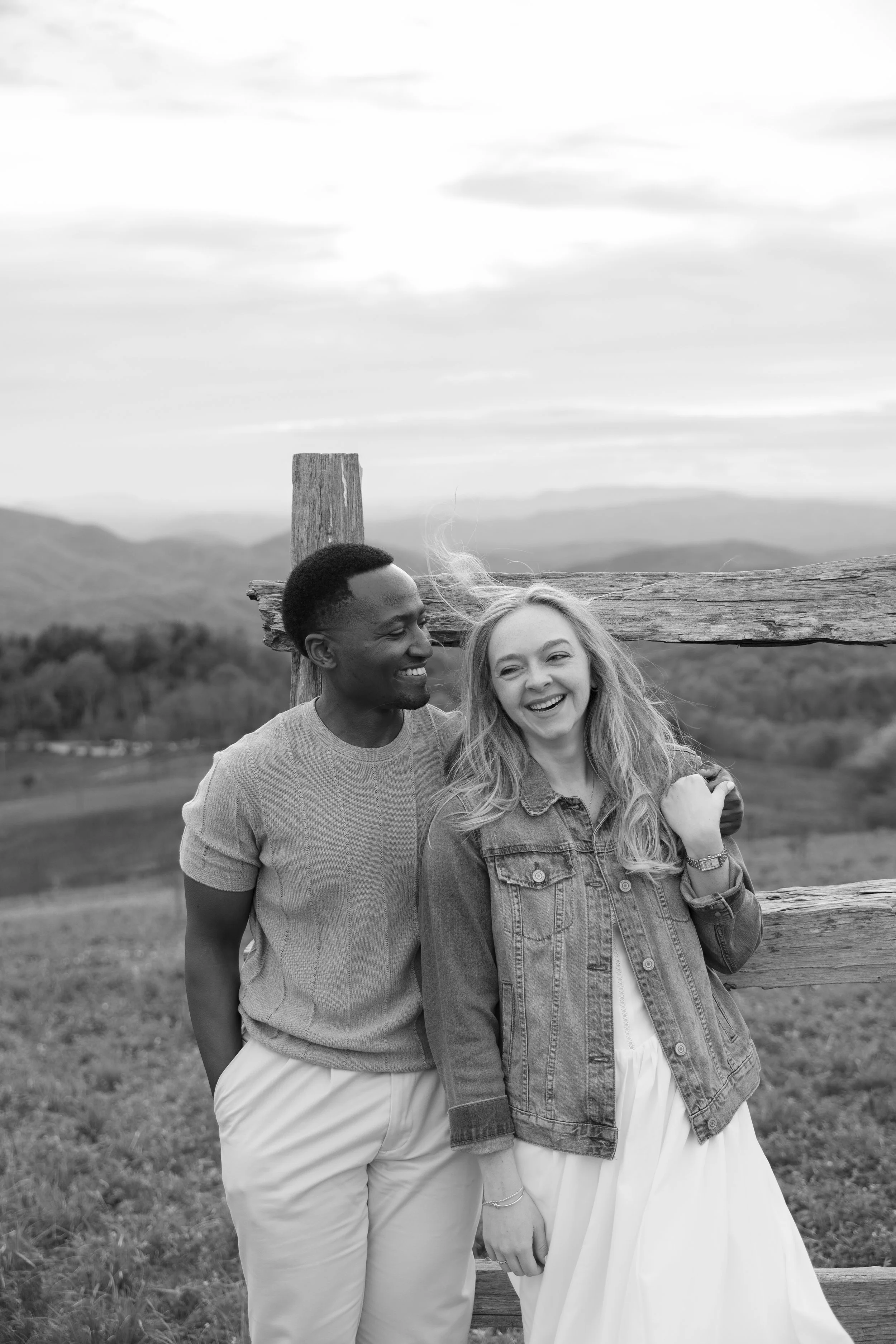 A black and white photo of a smiling couple standing outdoors in front of a wooden fence, with mountains and a cloudy sky in the background.