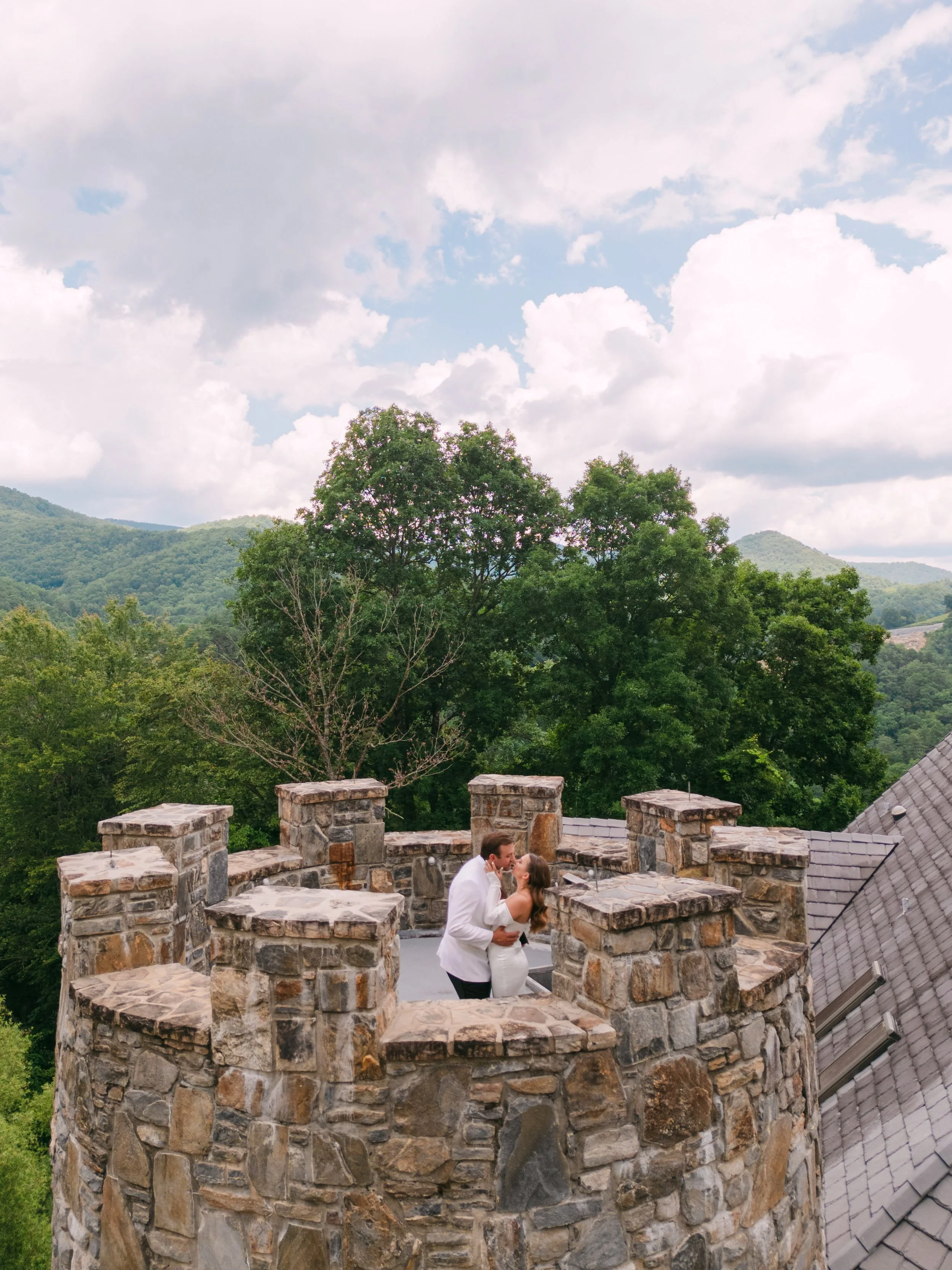 A bride and groom sharing a kiss on a stone tower with a scenic mountain and tree background.