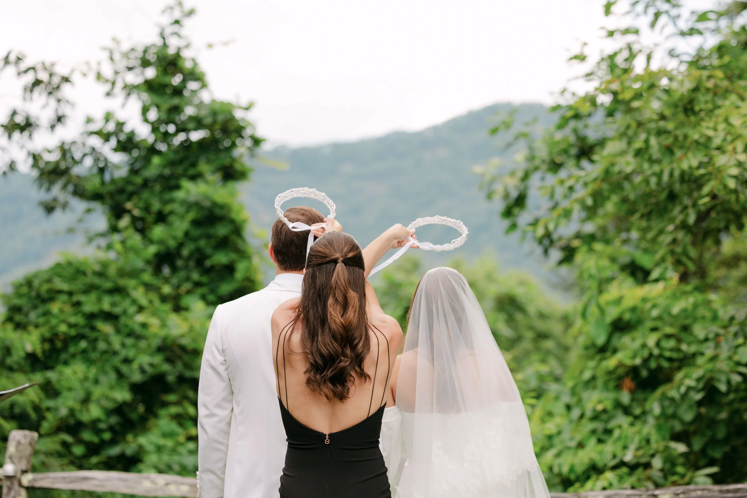 A bride, groom, and a woman with long brown hair in a black dress holding angel halo wands, standing outdoors with green trees and mountains in the background.