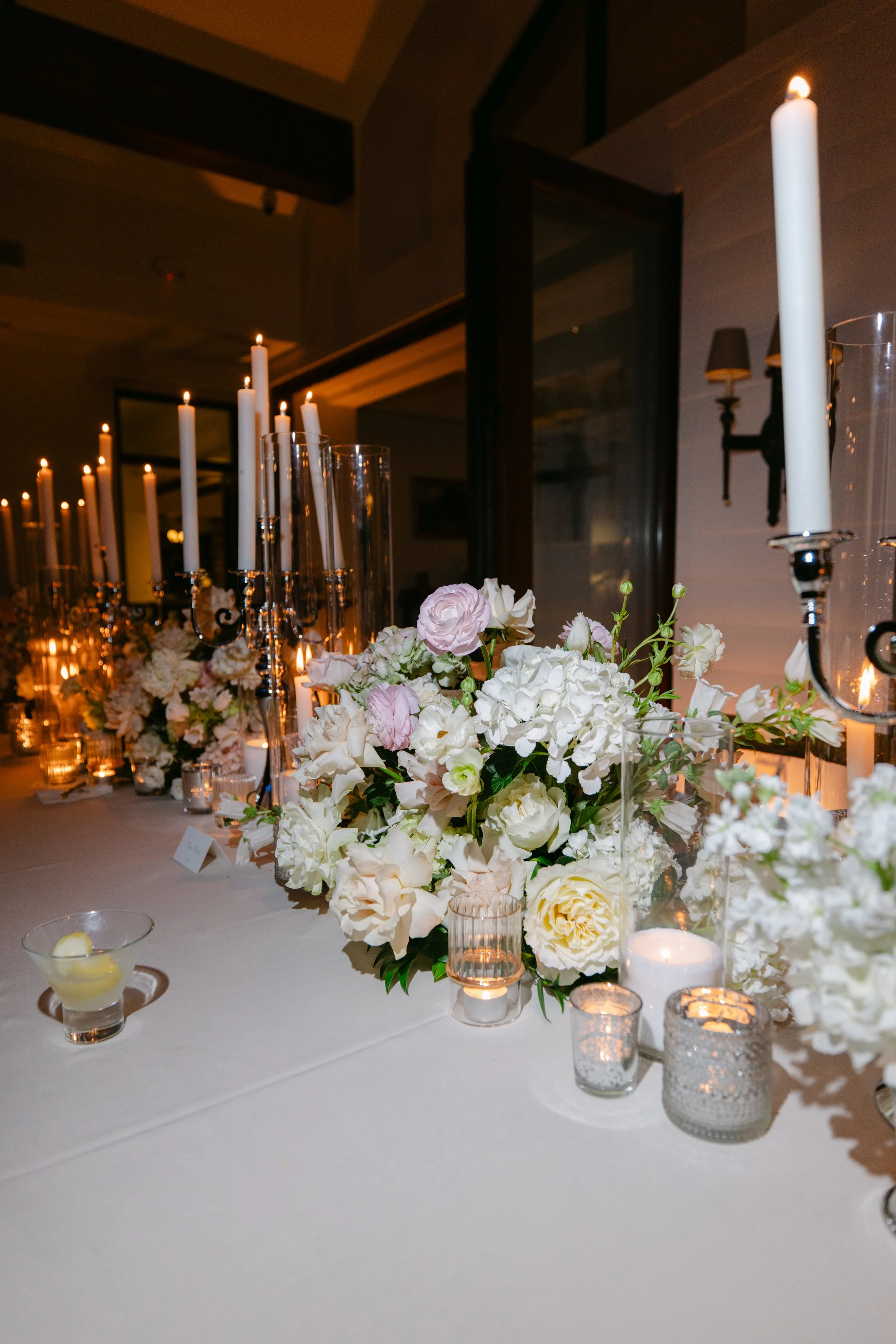 Elegant wedding reception table with white and blush floral arrangements, glowing candles, glass votive holders, and cocktail glasses