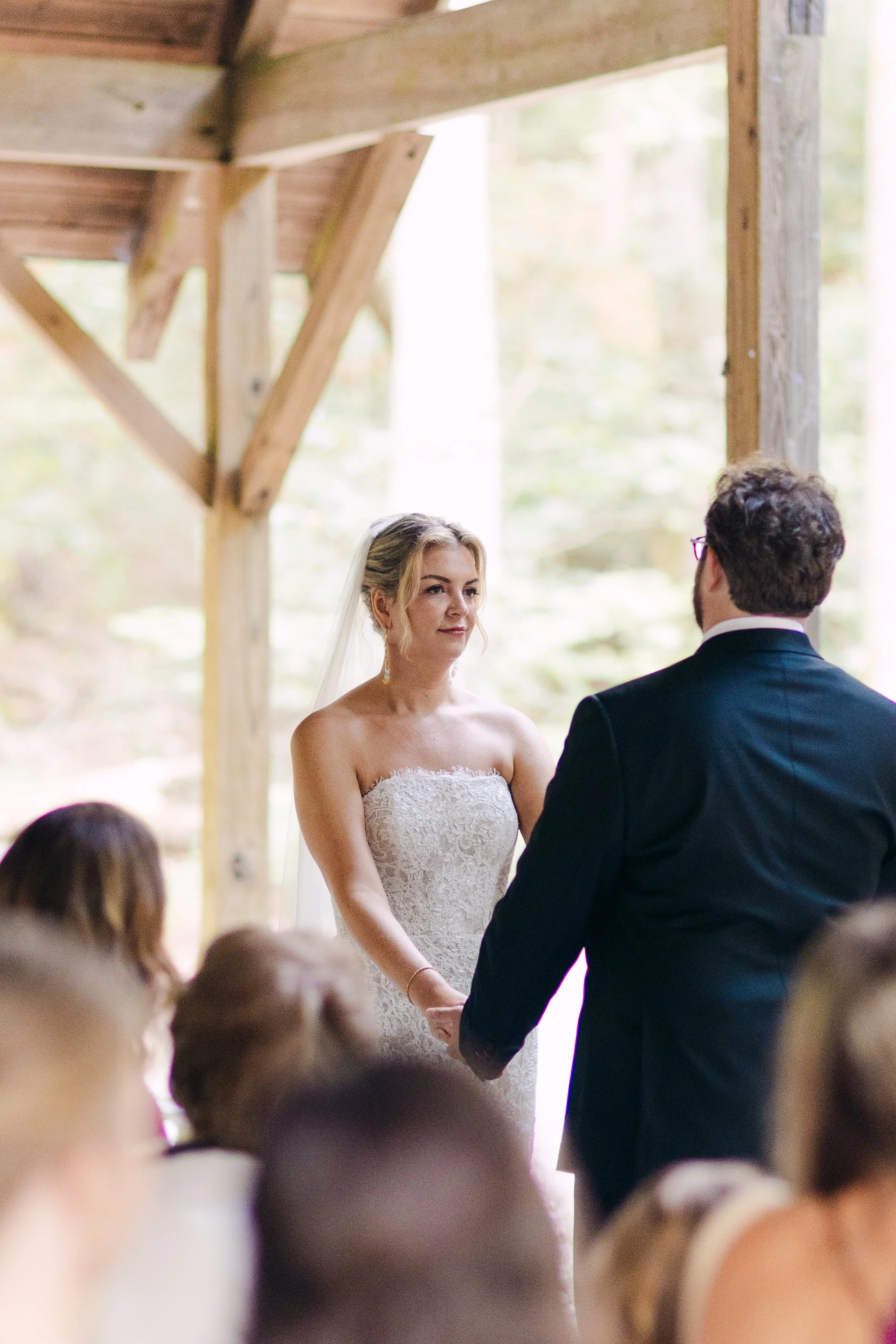 A bride and groom holding hands during their wedding ceremony in an outdoor wooden pavilion, with guests seated around them and trees in the background.