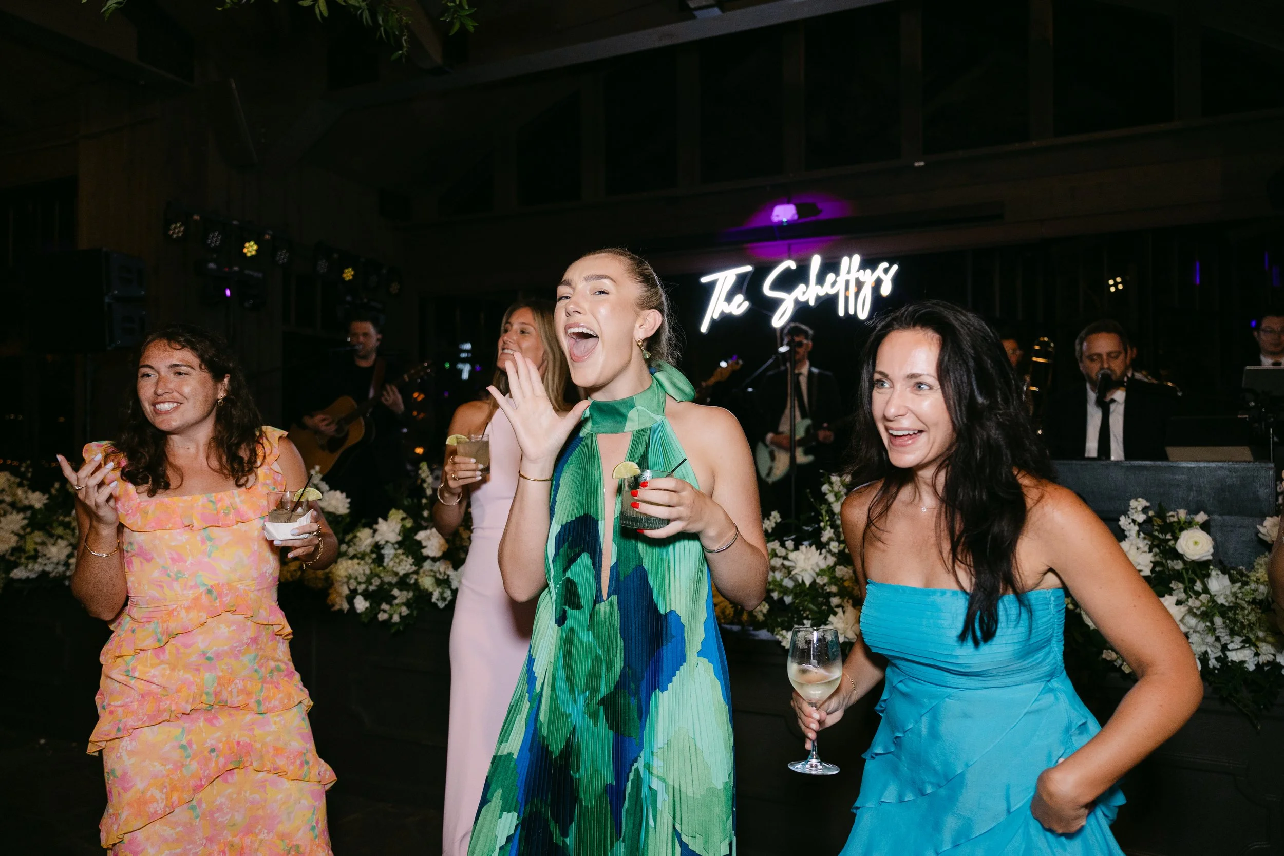 Four women in colorful dresses celebrating at a party, with a band playing live music in the background and a neon sign that reads 'The Shafty'.