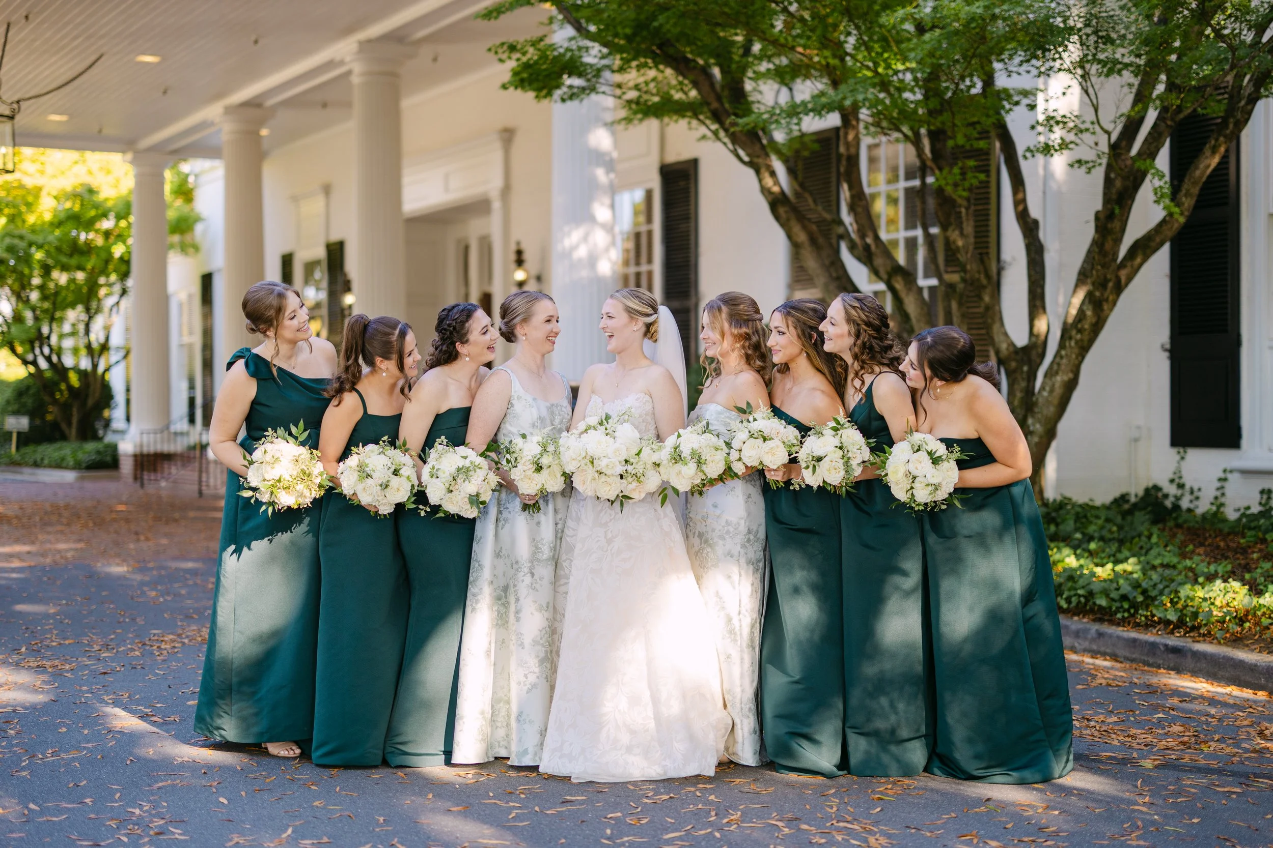 A group of women, including the bride and bridesmaids, standing outdoors on a street, smiling and holding bouquets of white flowers during a wedding celebration.