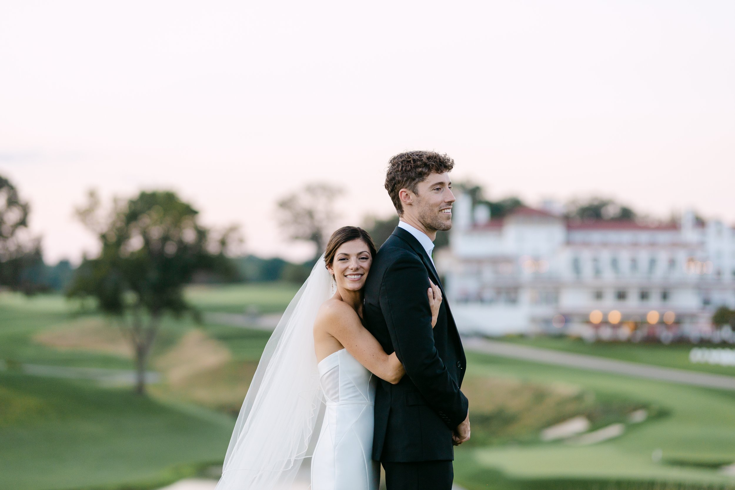 A newlywed couple standing outdoors on a golf course with a large building in the background. The bride is hugging the groom from behind, smiling happily.