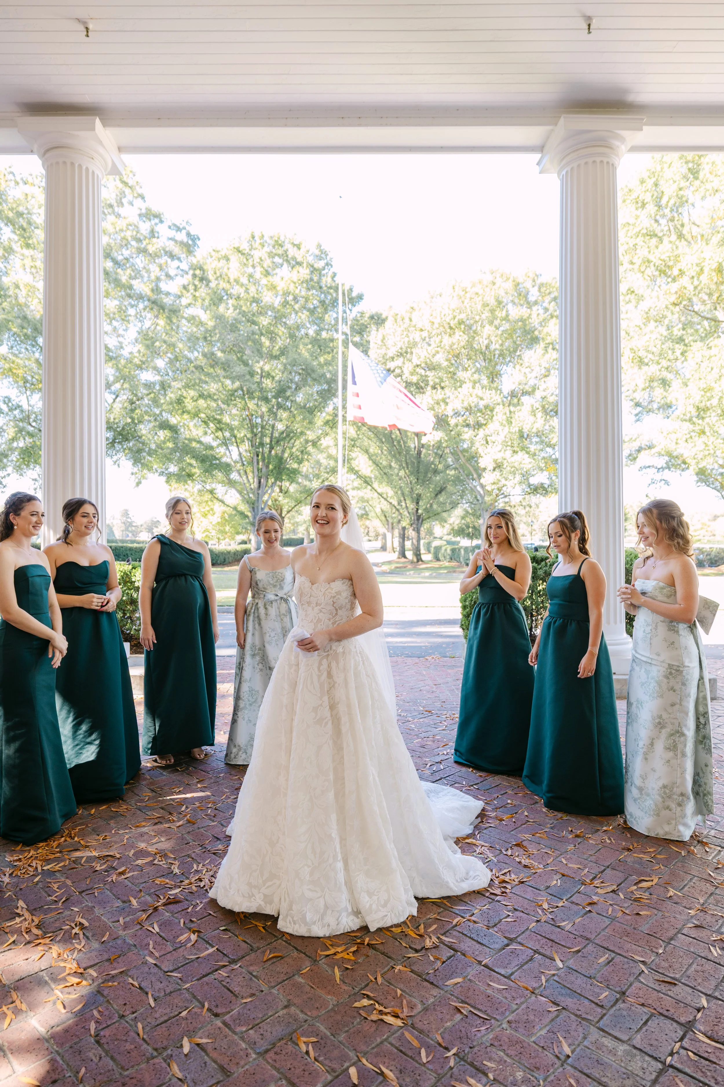 Bride in a white wedding gown smiling with bridesmaids in green dresses on the porch of a building with tall columns, autumn leaves on the brick ground, American flag in the background.