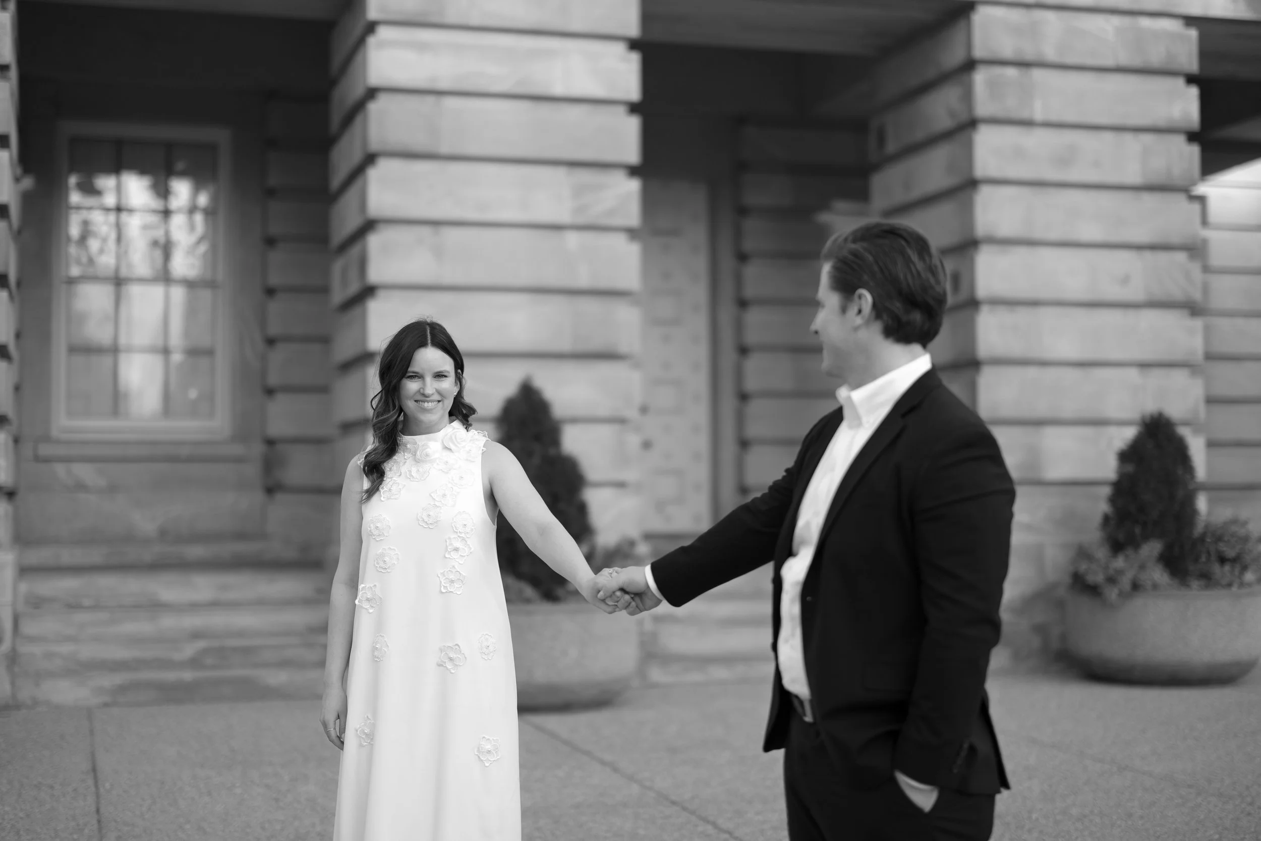 A woman in a white dress and a man in a black suit holding hands outside a building with large brick columns and potted plants.