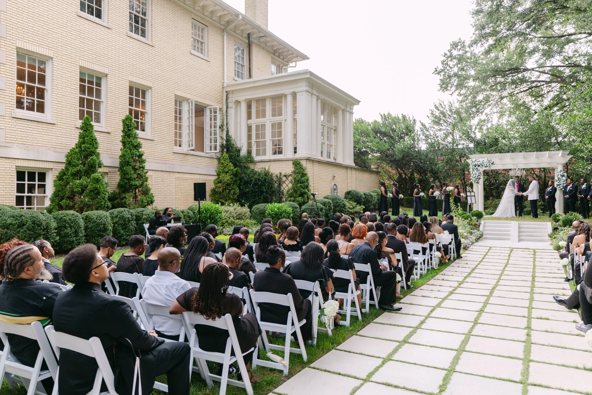 A wedding ceremony outdoors in the garden of a large historic building with cream bricks and white columns. The bride and groom stand under an arch with white flowers, facing each other as the officiant presides. Guests sit in white chairs, dressed f