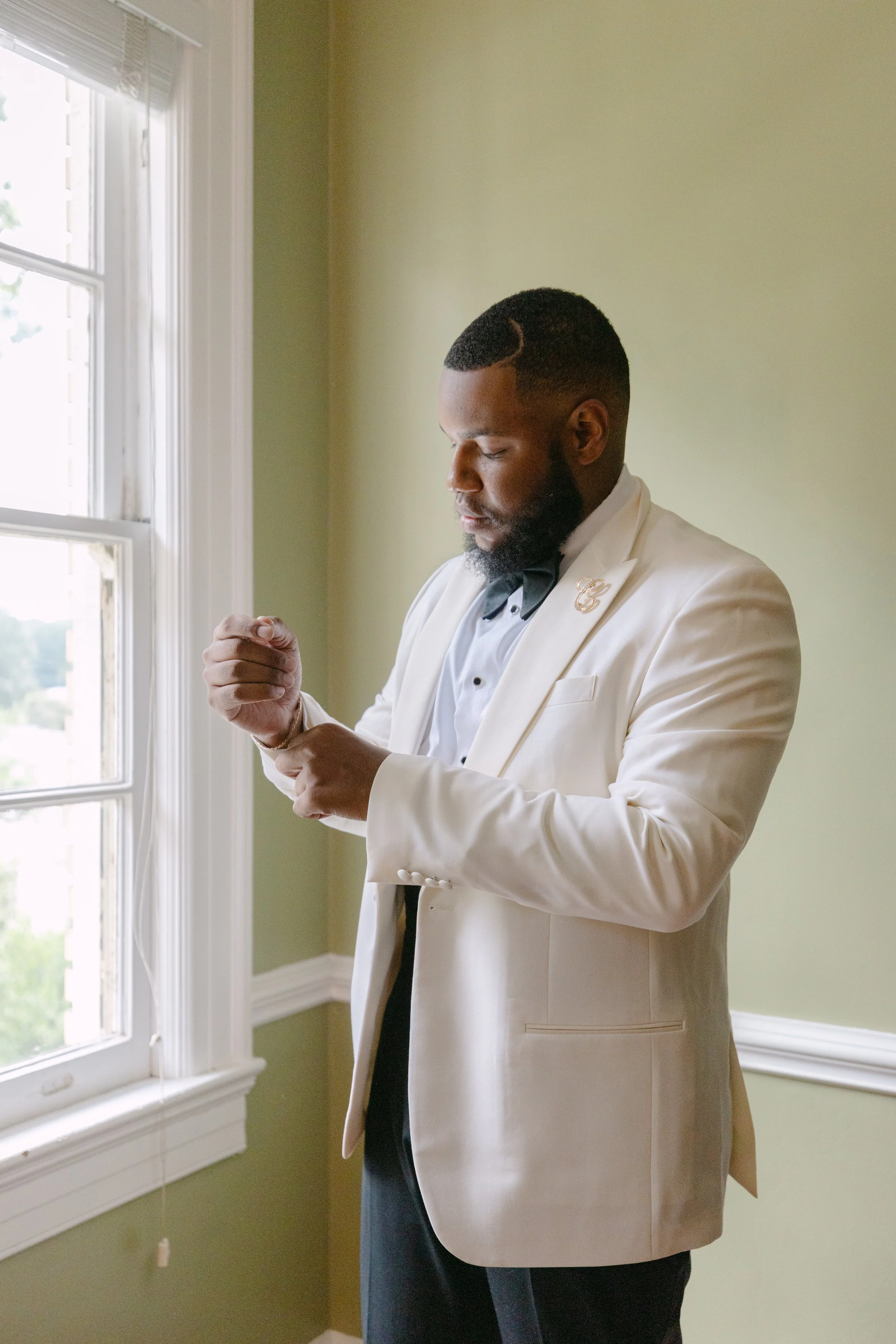 A man dressed in a white tuxedo with a black bow tie and a gold brooch, standing near a window with green walls, looking down and adjusting his cufflinks.