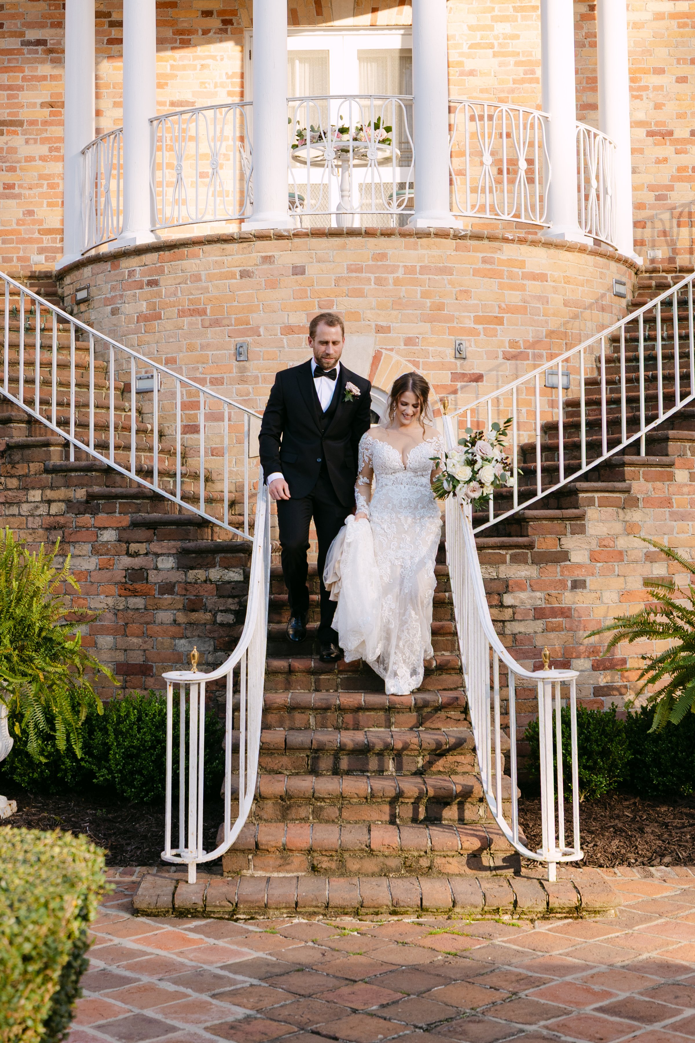 A bride and groom walking down brick stairs outside a brick building with a balcony, greenery on either side, during a wedding celebration.