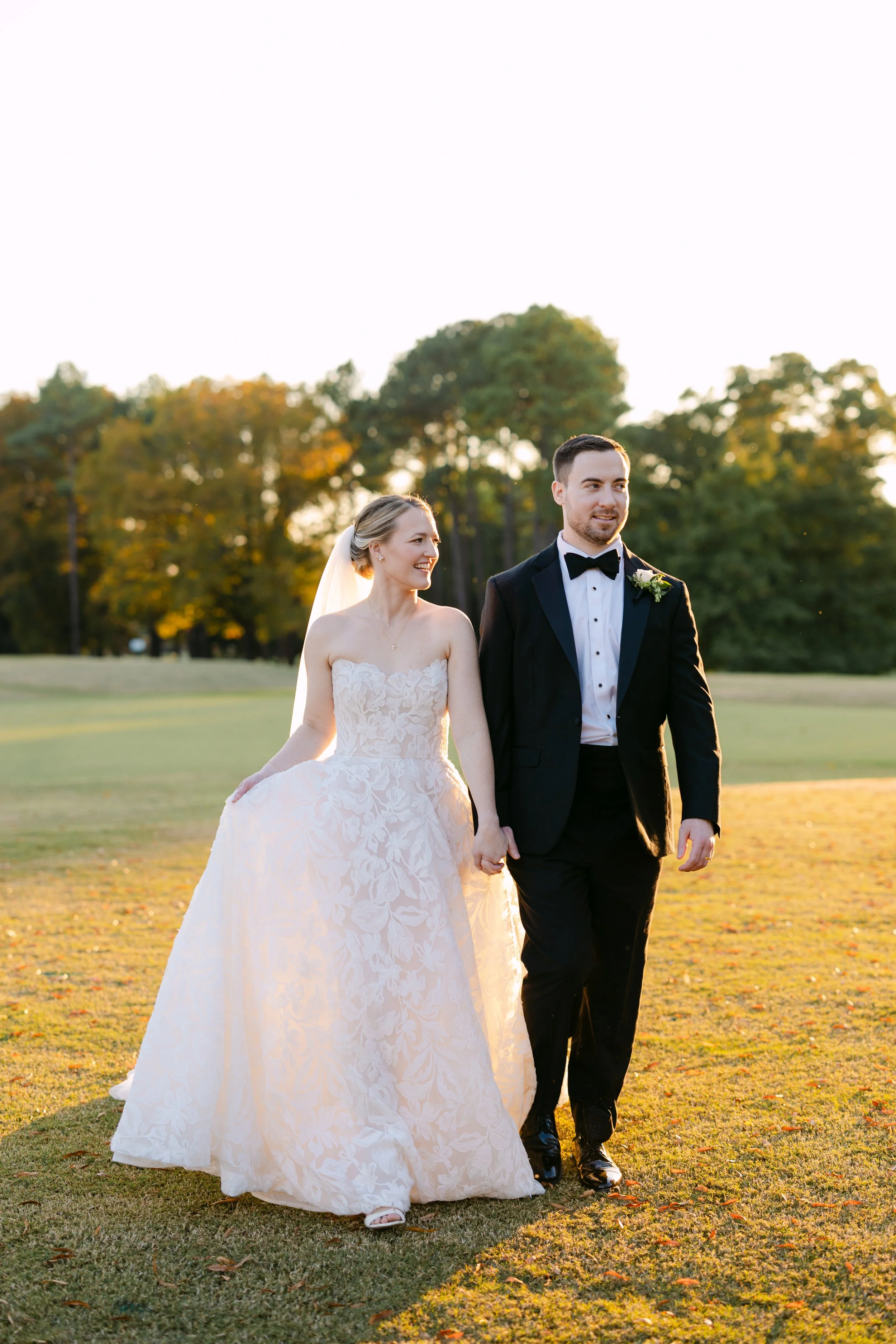 A bride and groom walking hand in hand on a grassy field during sunset, with trees in the background.