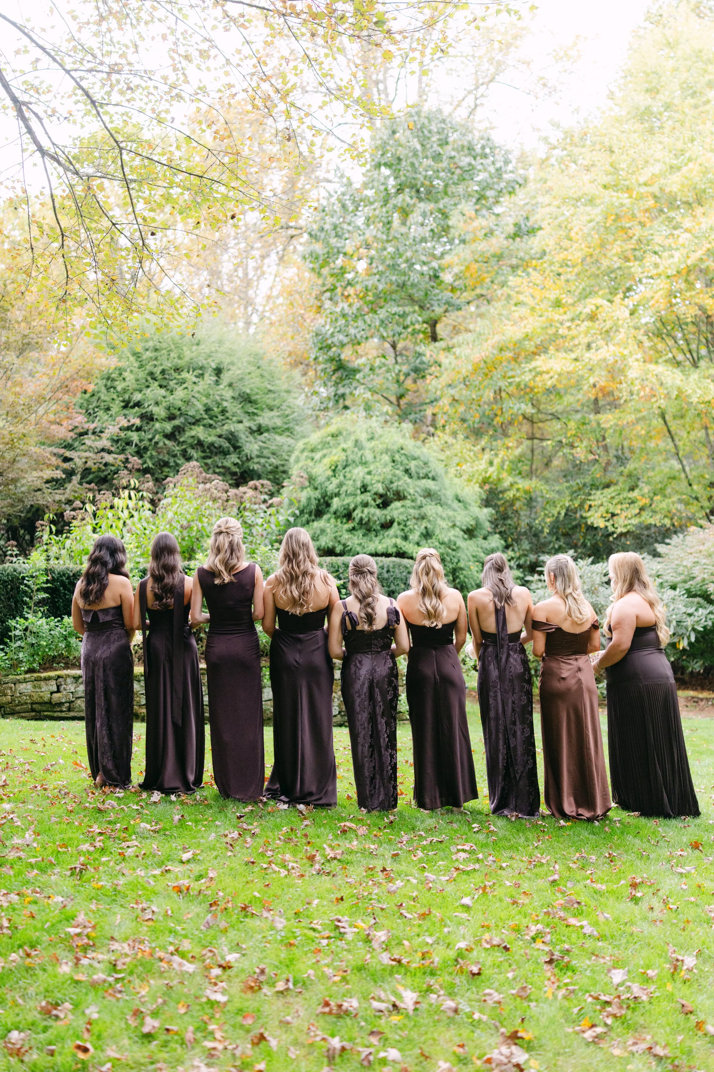 Group of women in dark formal dresses standing in an autumn garden surrounded by trees and fallen leaves