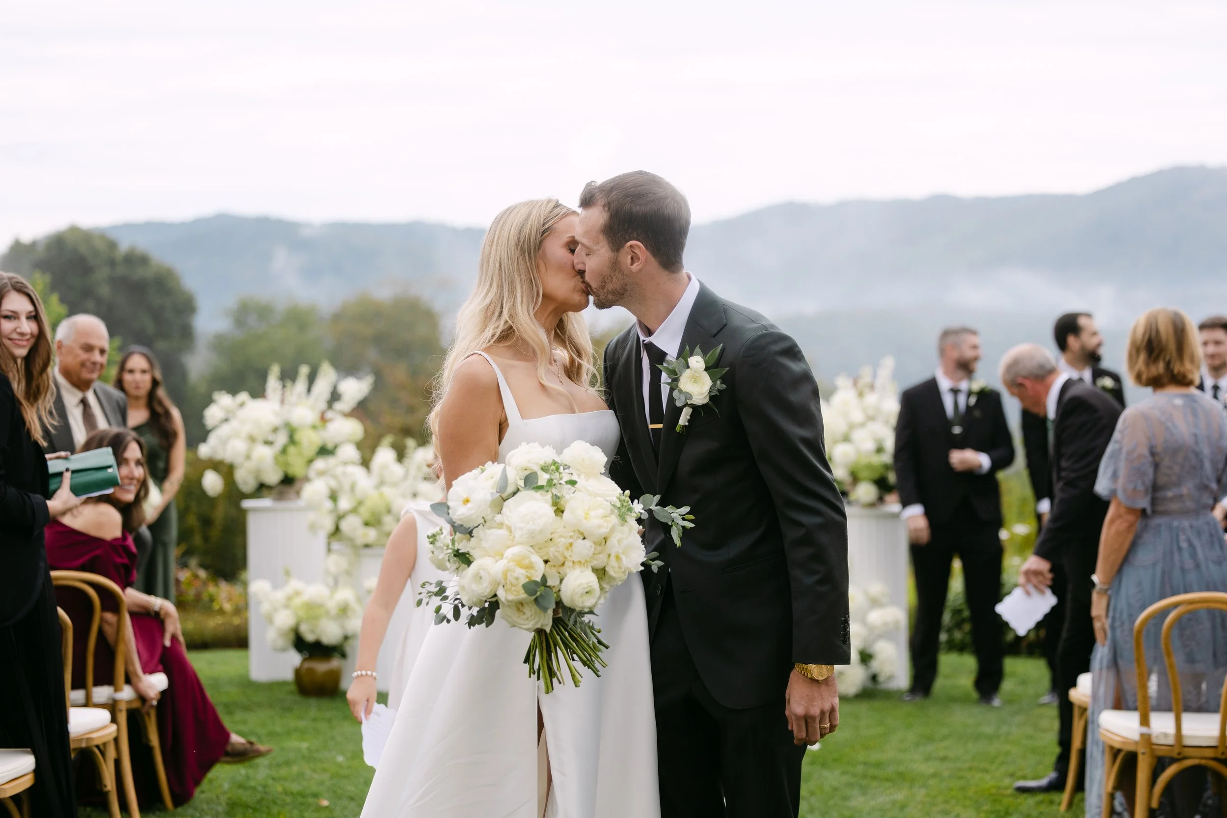 Couple in wedding attire sharing a kiss at an outdoor wedding ceremony, surrounded by friends and family, with white floral decorations and green landscape in the background.