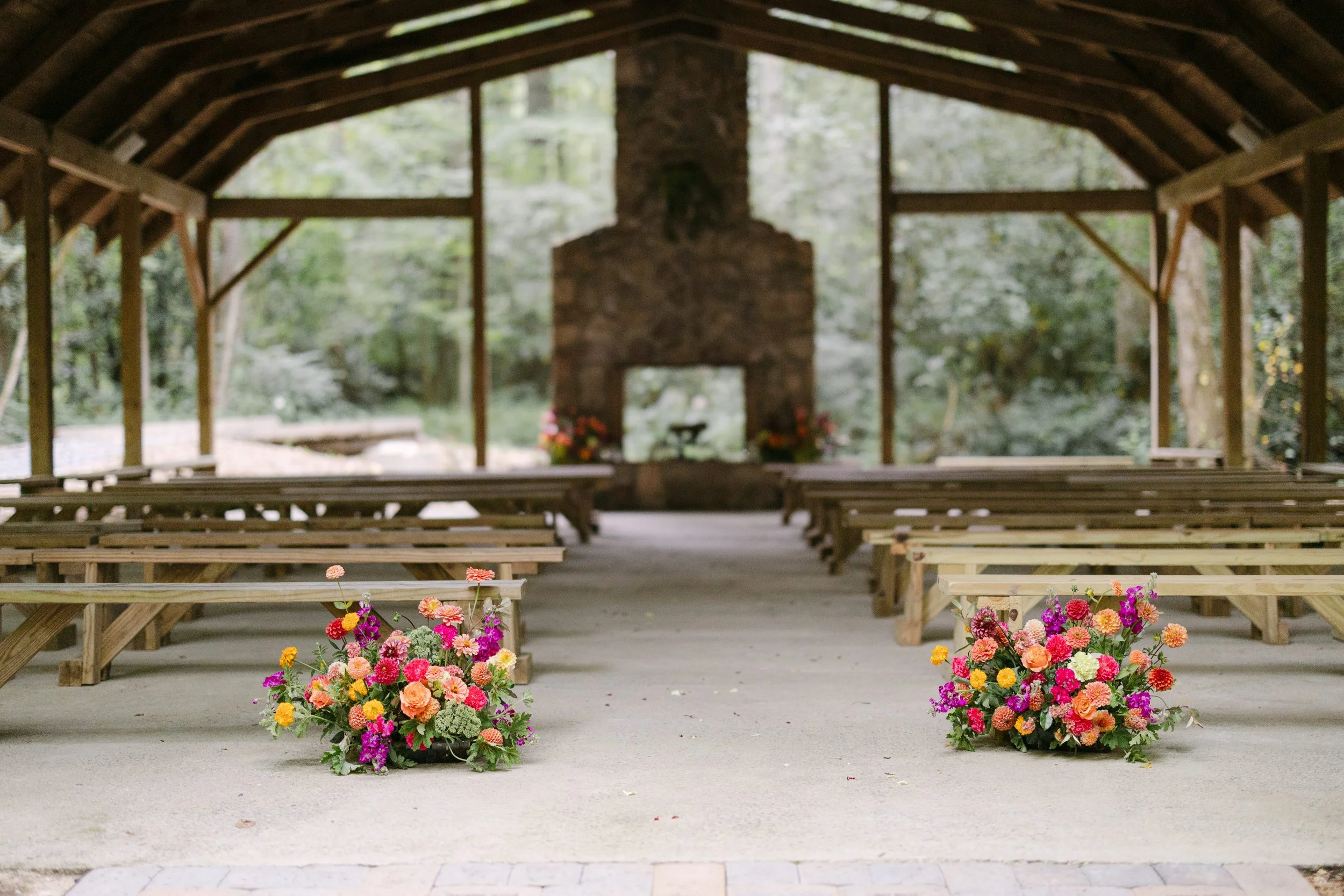 An outdoor covered pavilion with wooden benches and two large colorful flower arrangements on the ground, with a stone fireplace in the background decorated with flowers.