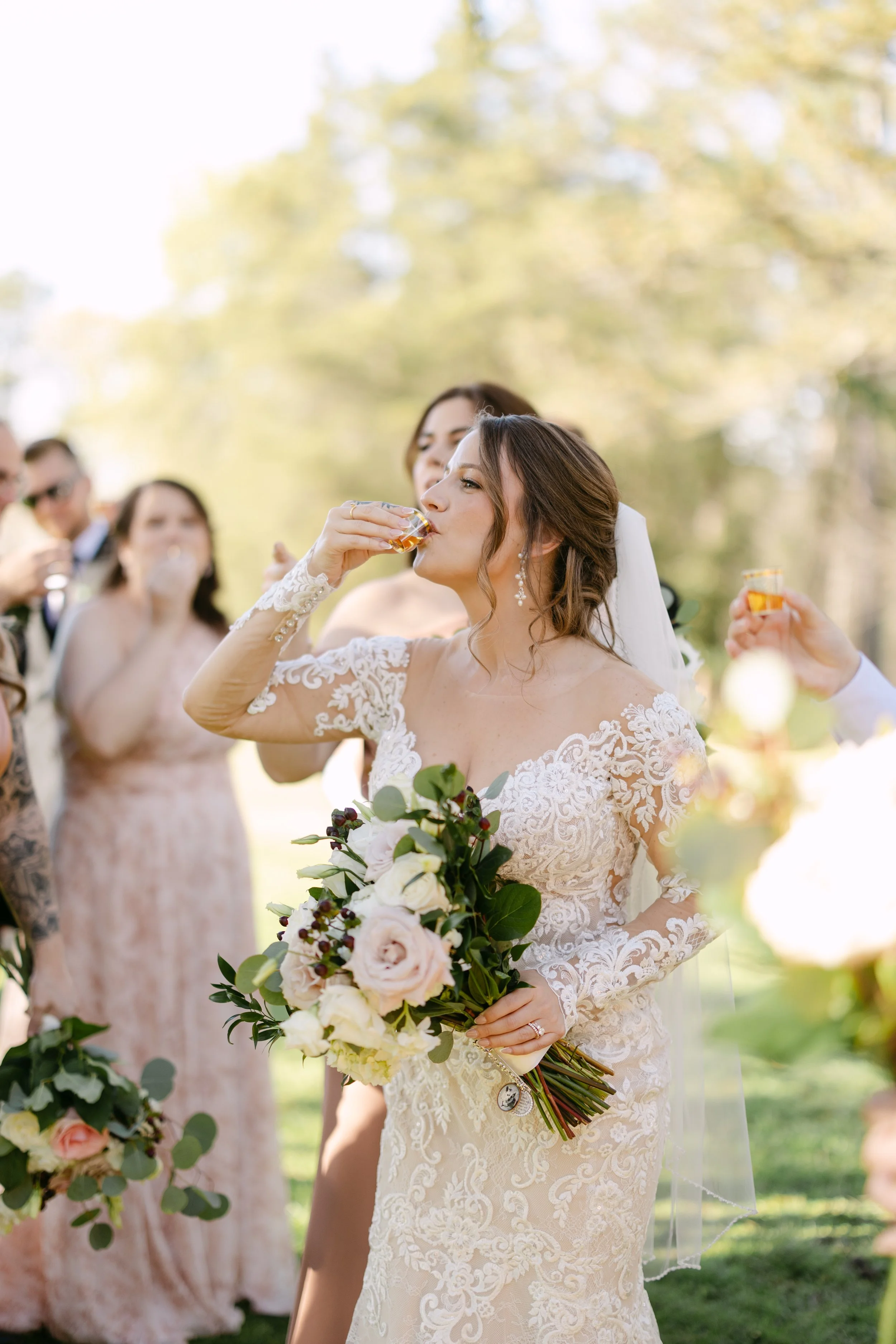 Bride in lace wedding dress toasting with friends outdoors during a wedding celebration.