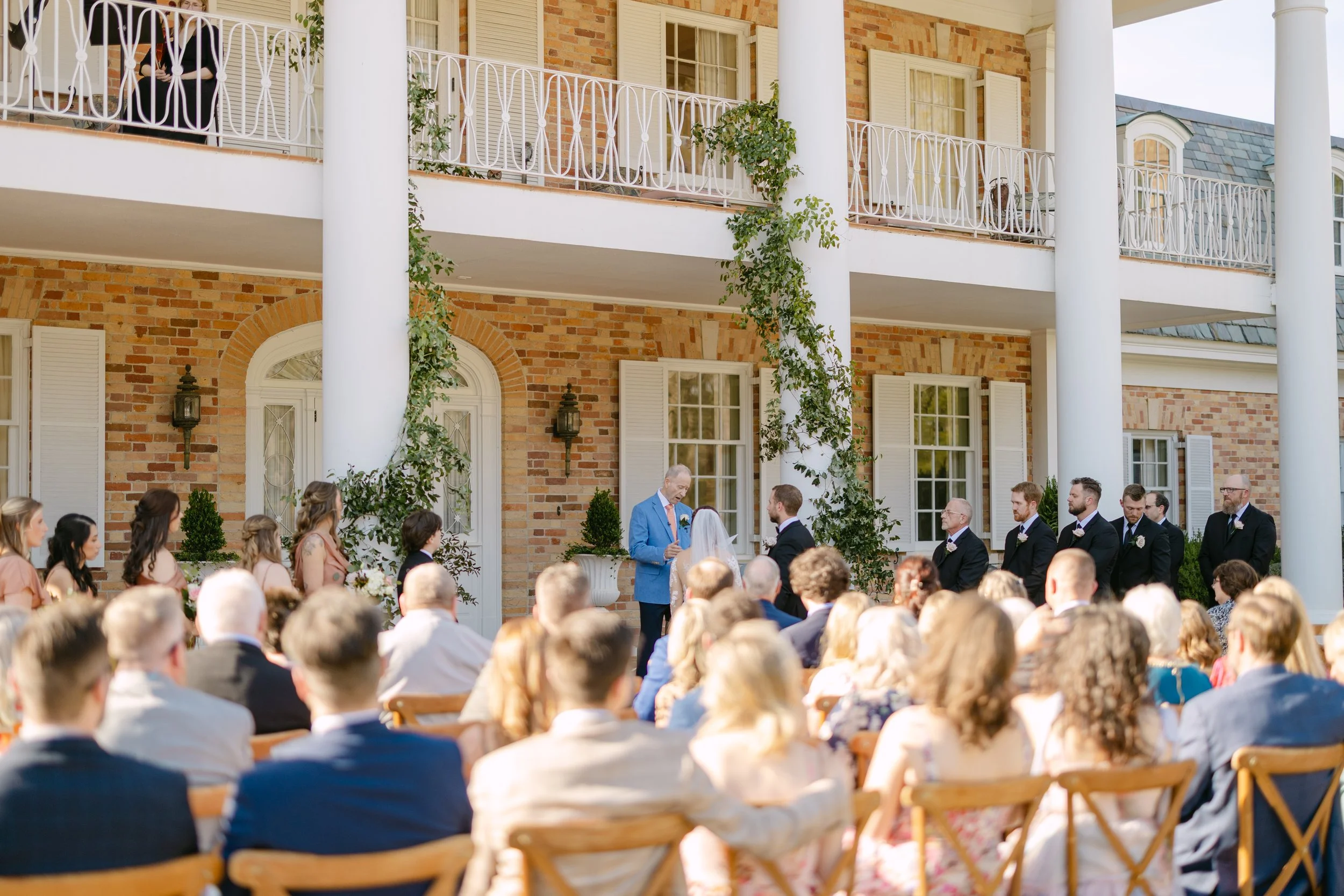 Outdoor wedding ceremony with a bride and groom exchanging vows, officiant, bridesmaids, and groomsmen, in front of a large brick building with columns and greenery, guests seated watching.