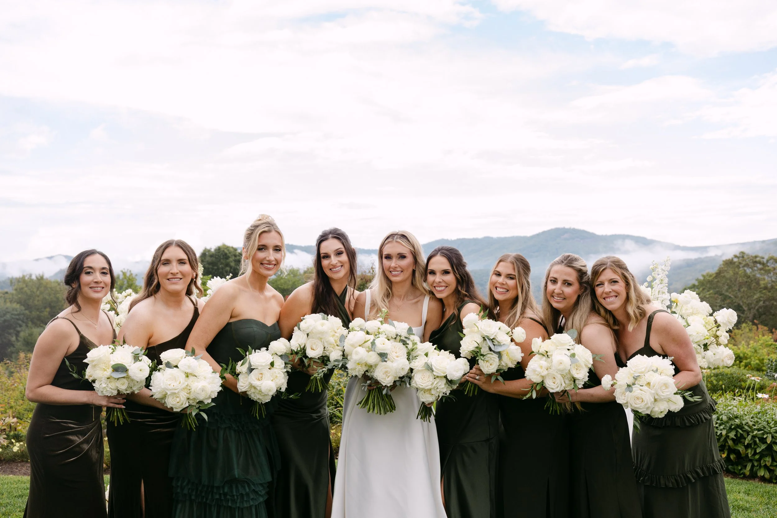 A group of nine women standing outdoors, dressed in black dresses, holding white bouquets, with a scenic mountain background and cloudy sky.