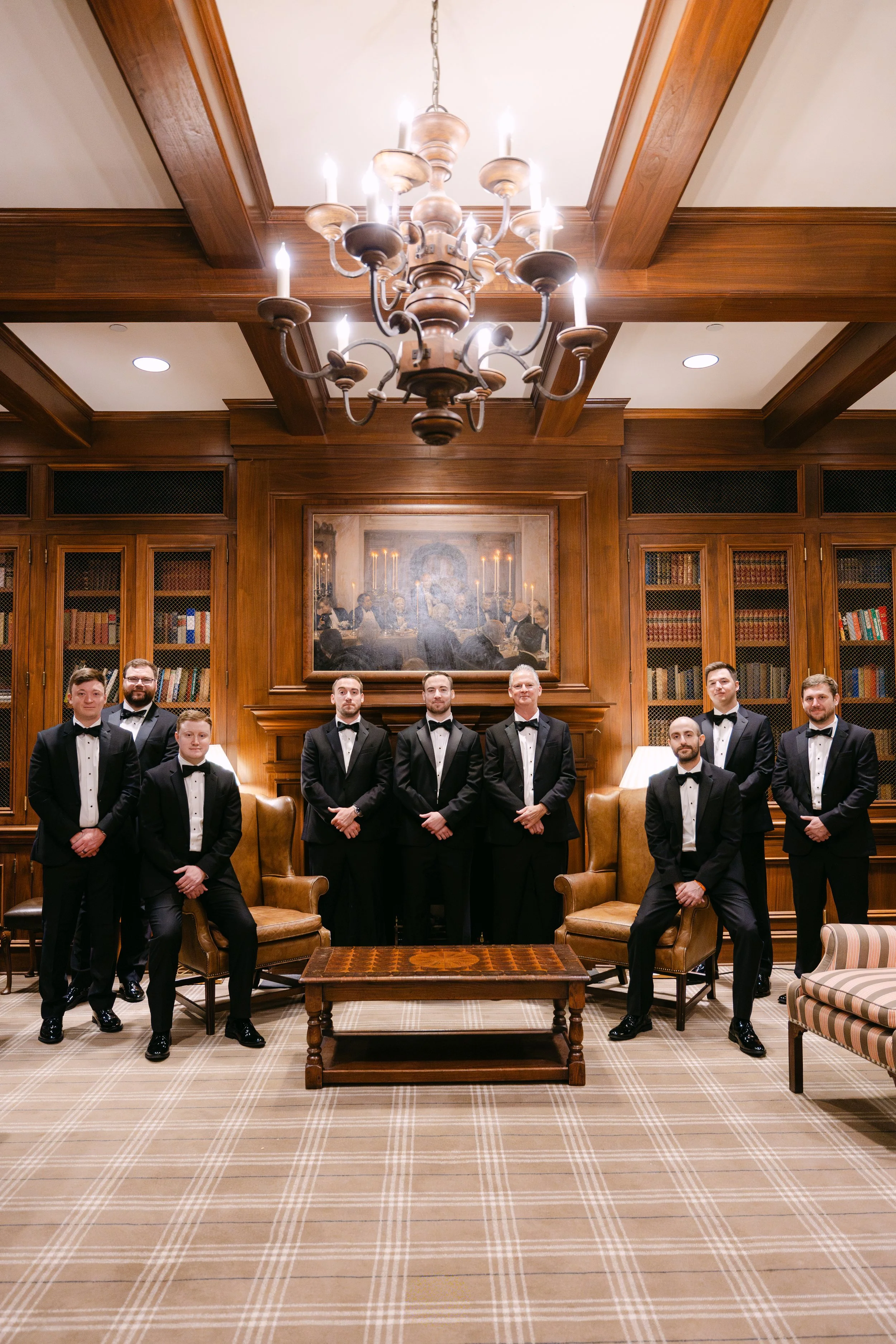 A group of nine men dressed in black tuxedos with bow ties, posing for a photo in a wood-paneled room with a chandelier, bookshelves, and leather chairs.
