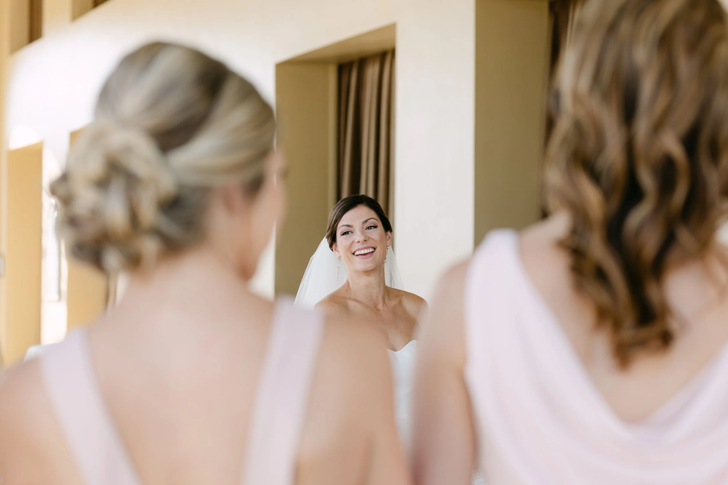 Bridal preparation scene with a bride looking at herself in a mirror while two bridesmaids watch, all dressed in white gowns.