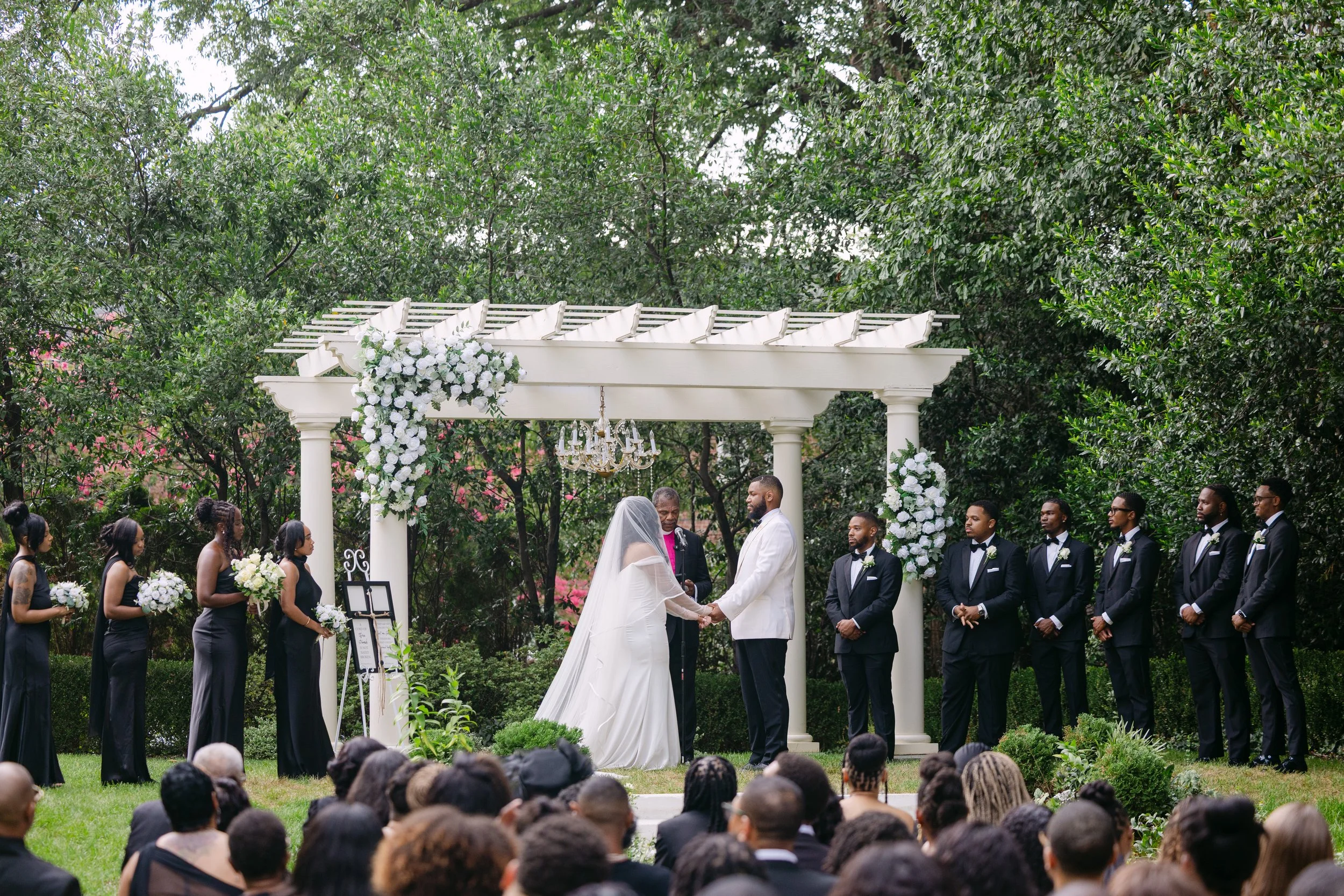 A wedding ceremony taking place outdoors under a white pergola decorated with white flowers, with the bride and groom holding hands and the wedding party standing on either side.