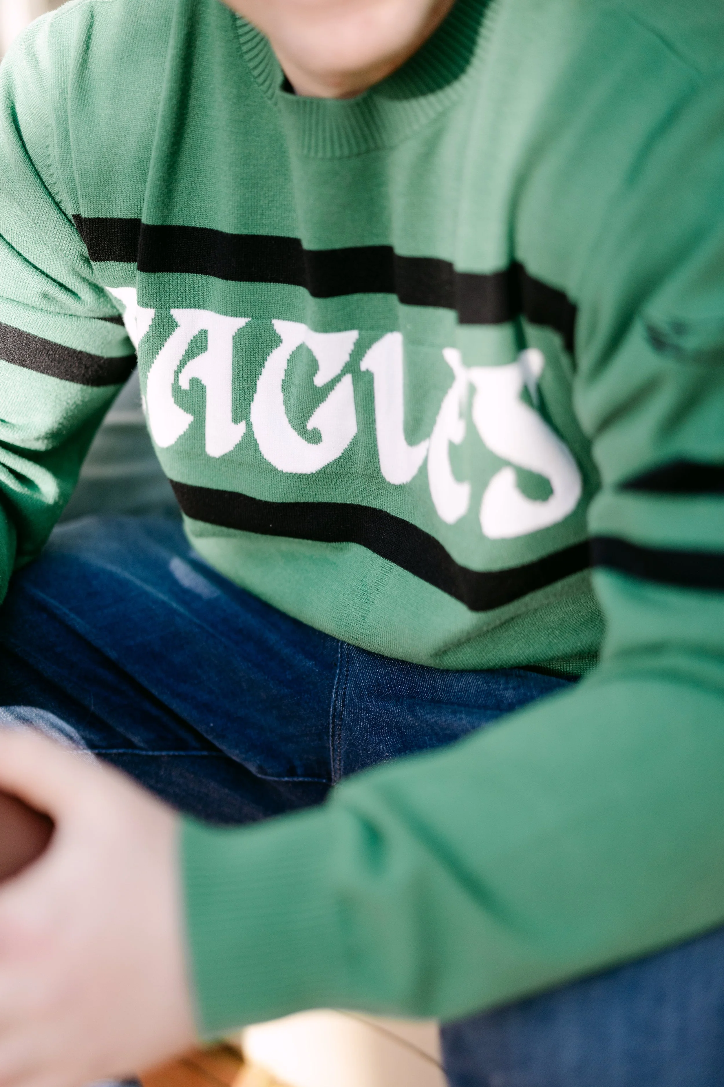 Close-up of a person wearing a green sweatshirt with black stripes and white text, sitting with knees bent, in a casual setting.