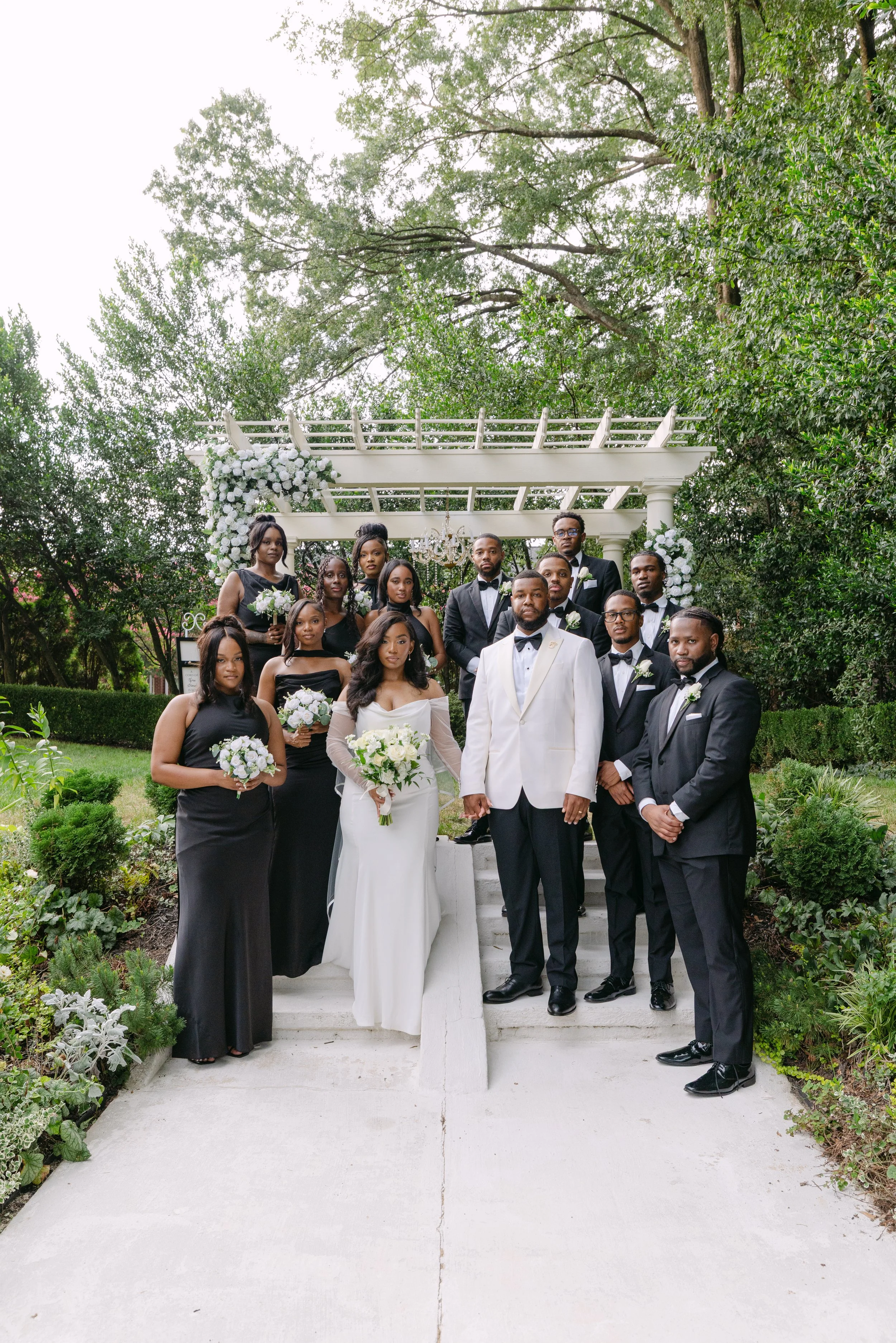 Group of people dressed in formal attire at a wedding, standing outdoors on steps with a white floral arch and greenery in the background.