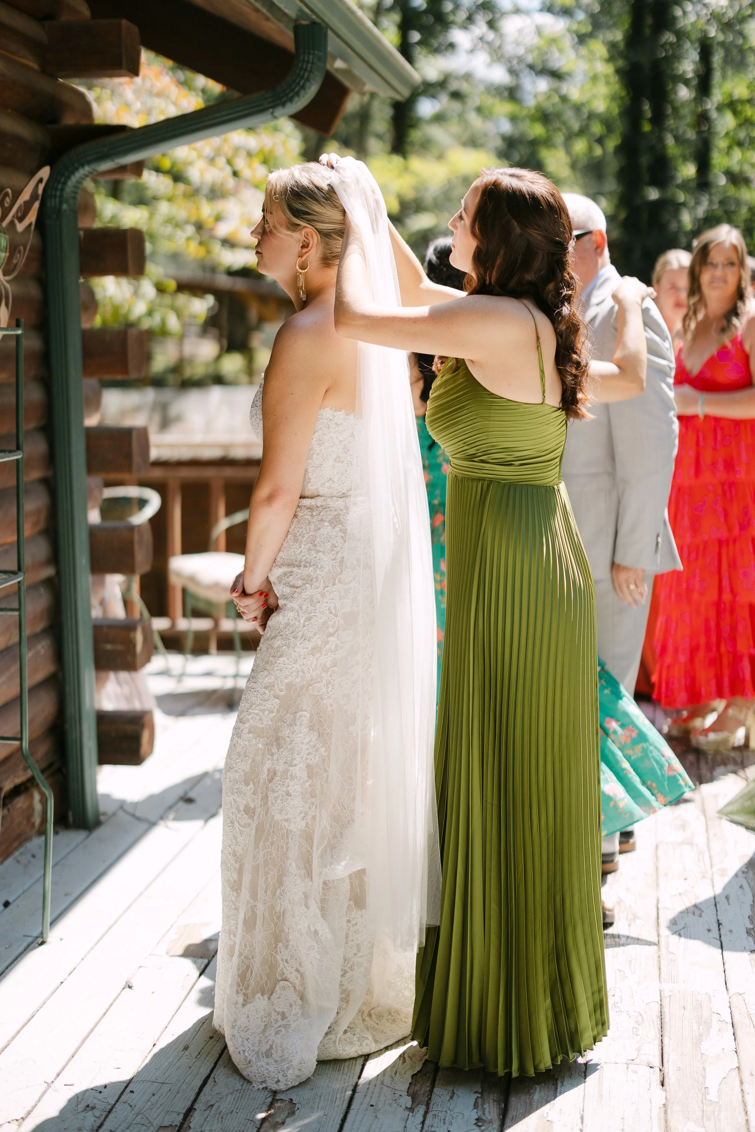 A woman in a white lace wedding dress getting her veil adjusted by another woman in a green pleated dress outdoors during a wedding ceremony, with guests in the background.