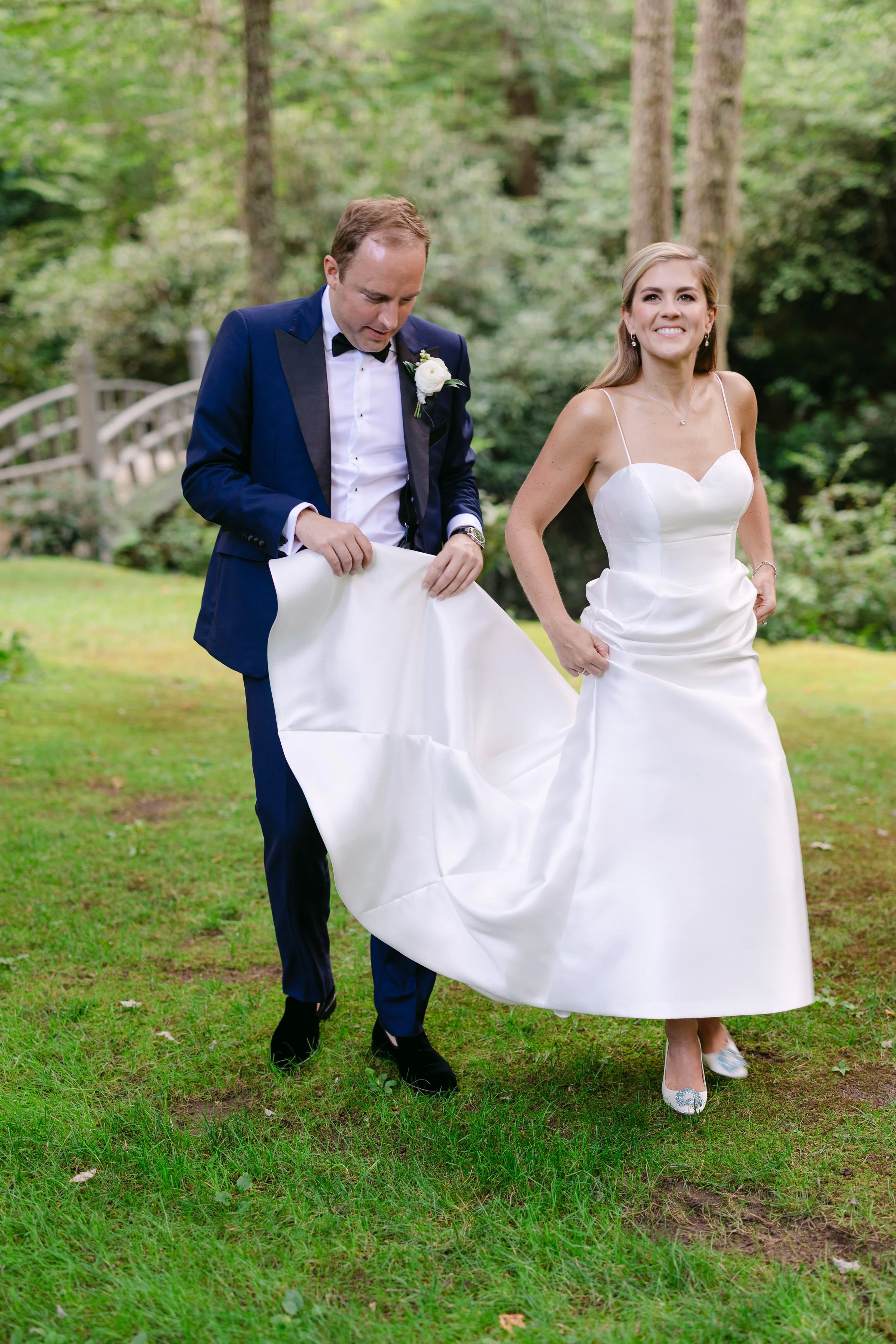 A bride and groom outdoors in a forested area, with the groom helping to lift the bride's wedding dress.