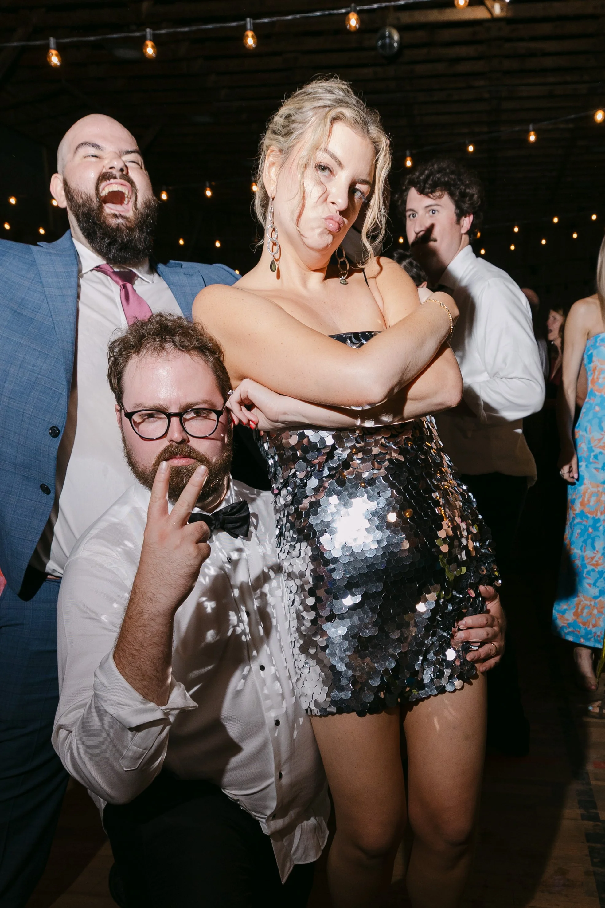 Group of people at a party wearing formal attire, with string lights overhead. One woman stands with crossed arms and a playful expression, dressed in a sequined dress. A man kneels in front making a peace sign, wearing glasses and a tuxedo. Two othe