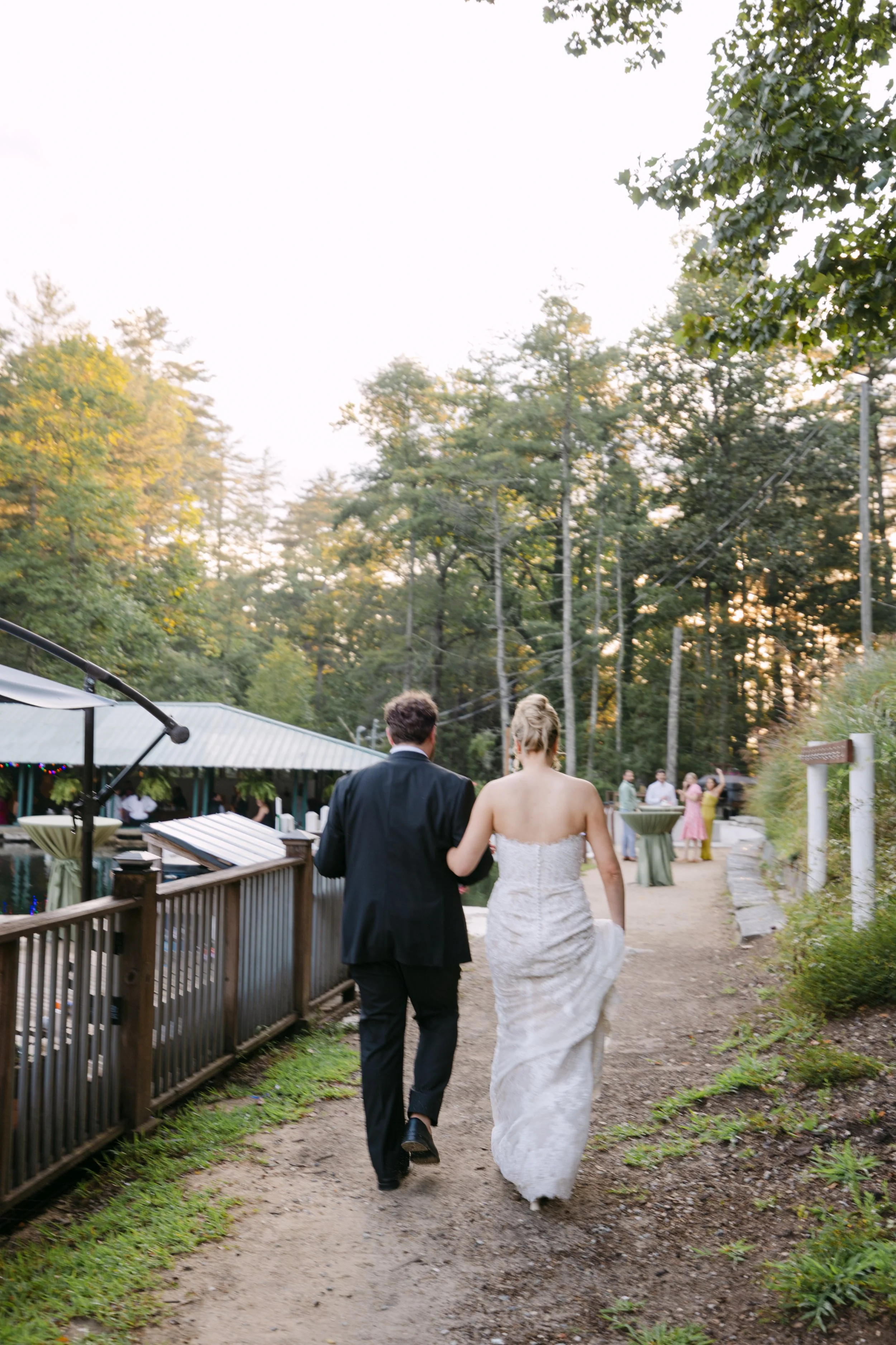 A couple in wedding attire walking along a dirt path at an outdoor reception area with trees and guests in the background.