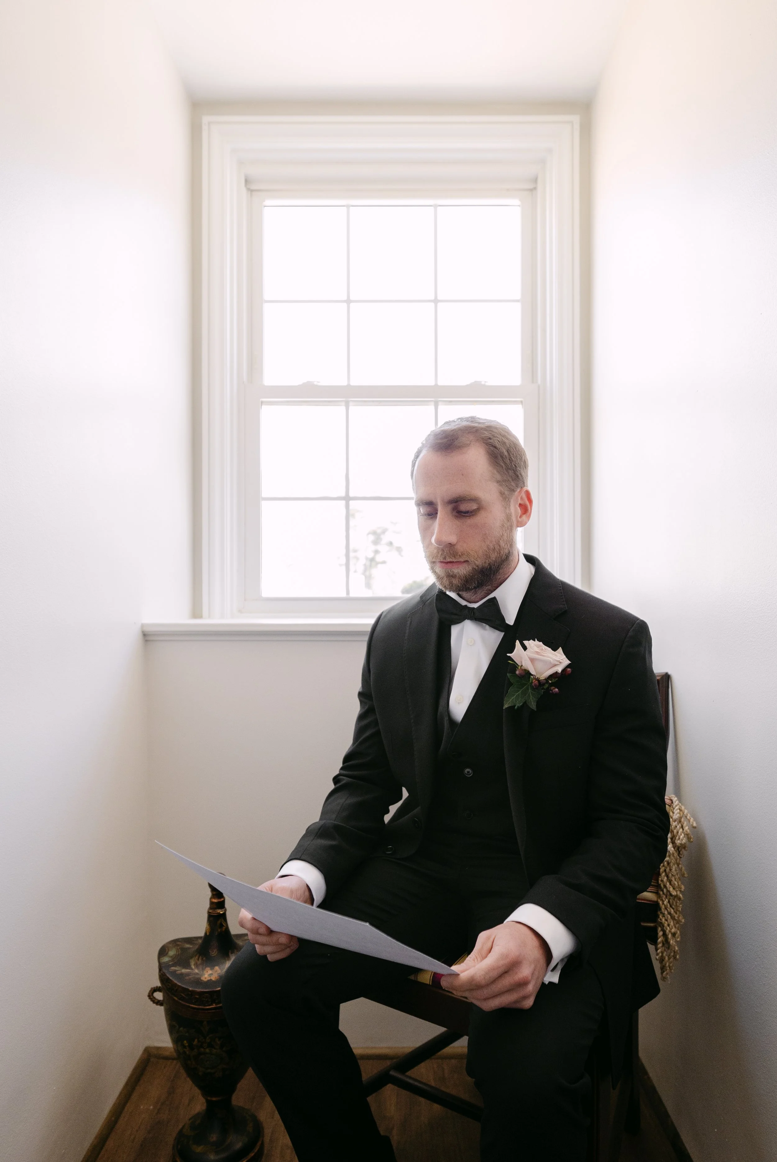A man in a black tuxedo with a boutonniere sits in a small, white-walled room near a window, reading a document.
