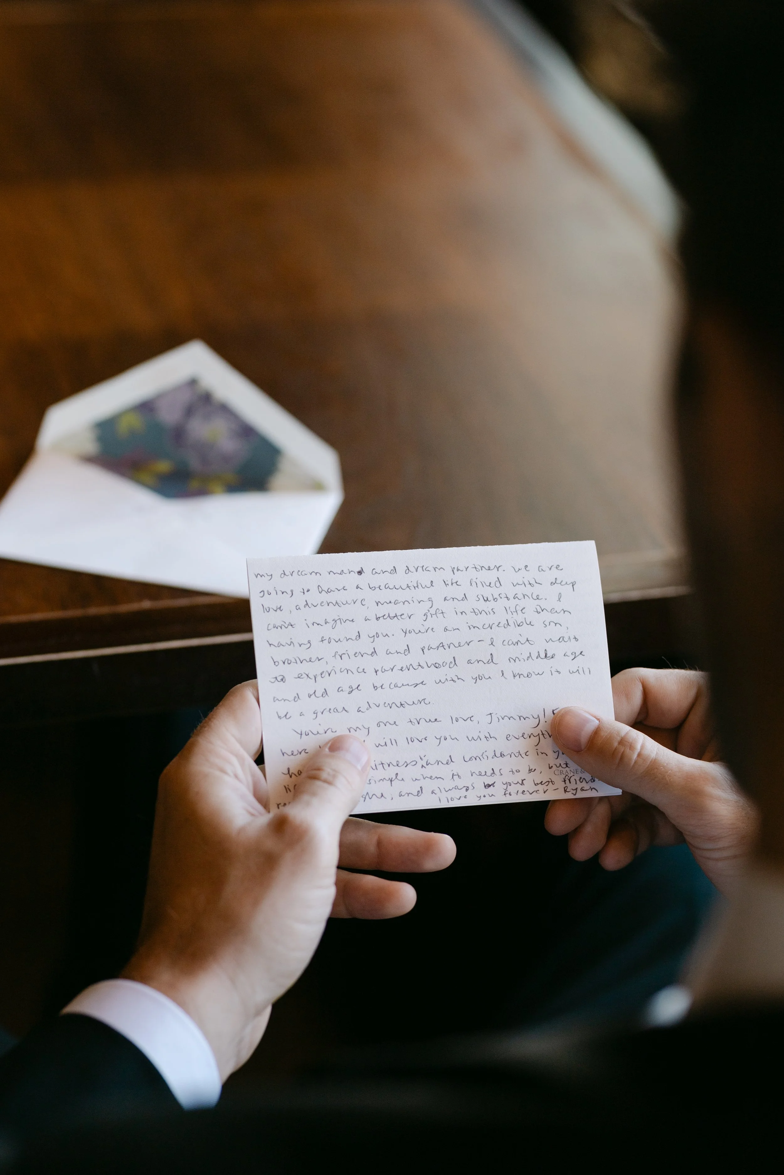 Person in black suit holding handwritten love letter with an envelope on a wooden surface.