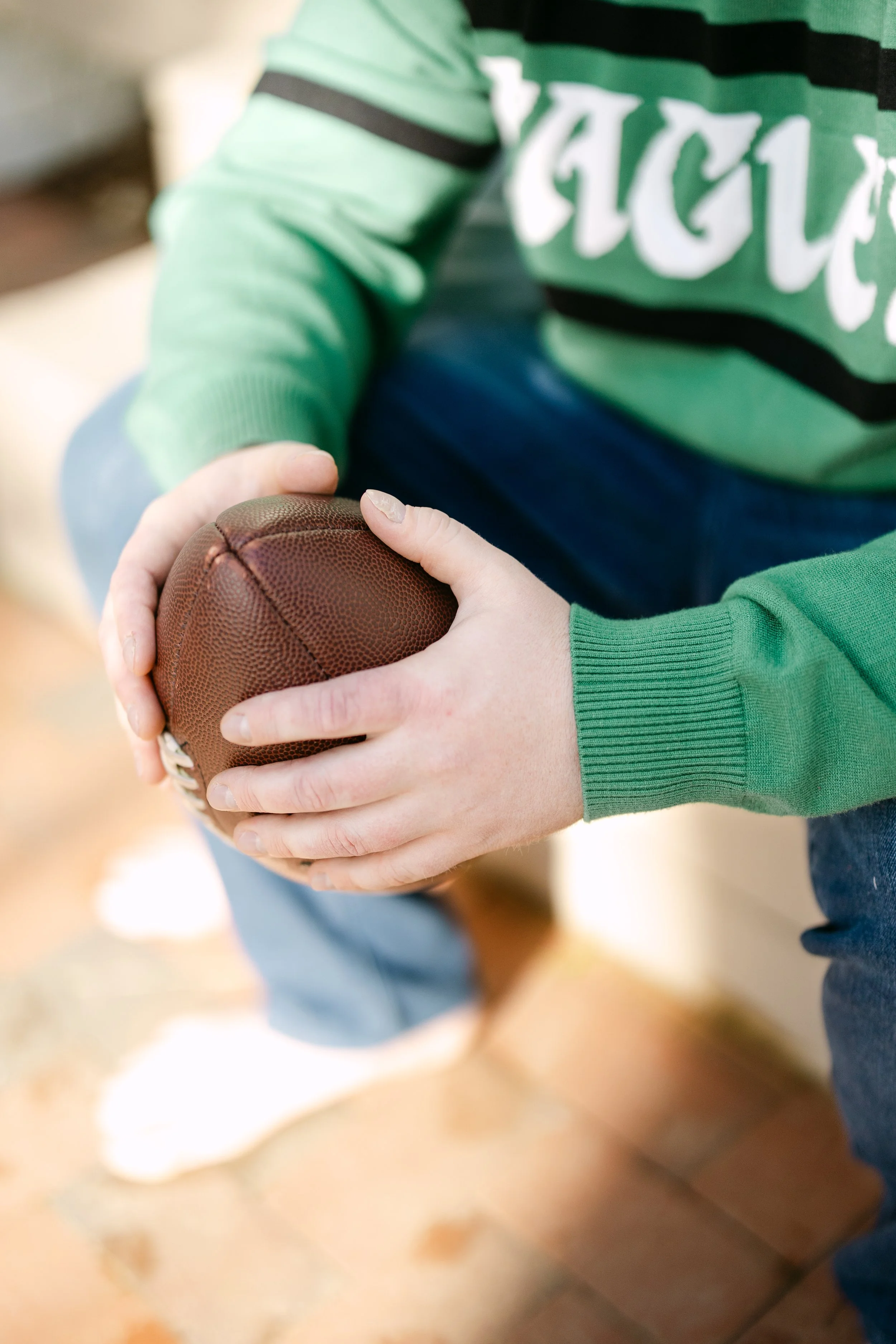 A person in a green and black hoodie holding a brown American football with both hands.