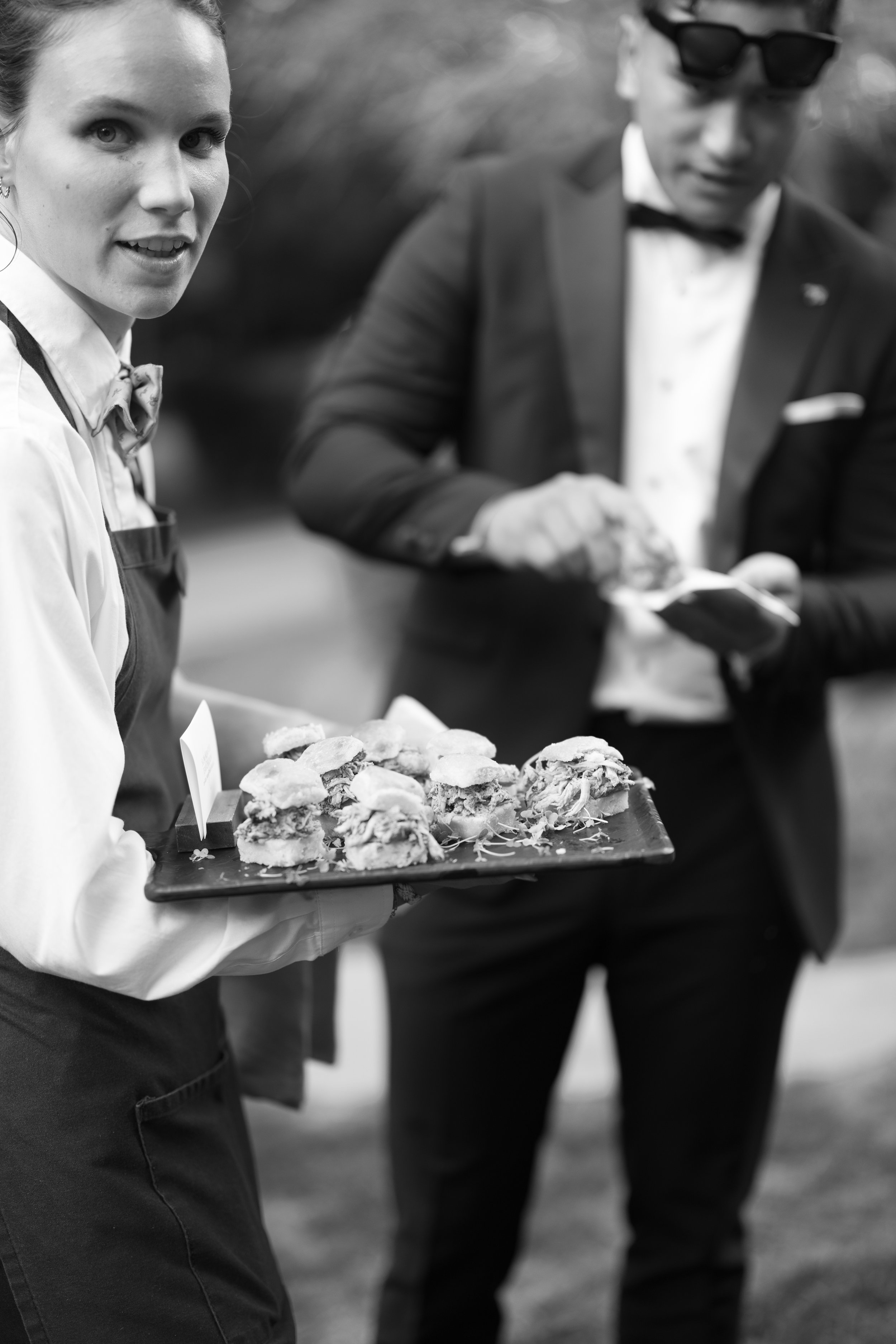 A woman in a white shirt and apron holding a tray of sandwiches, with a man in a suit and sunglasses using a phone in the background, outdoors.