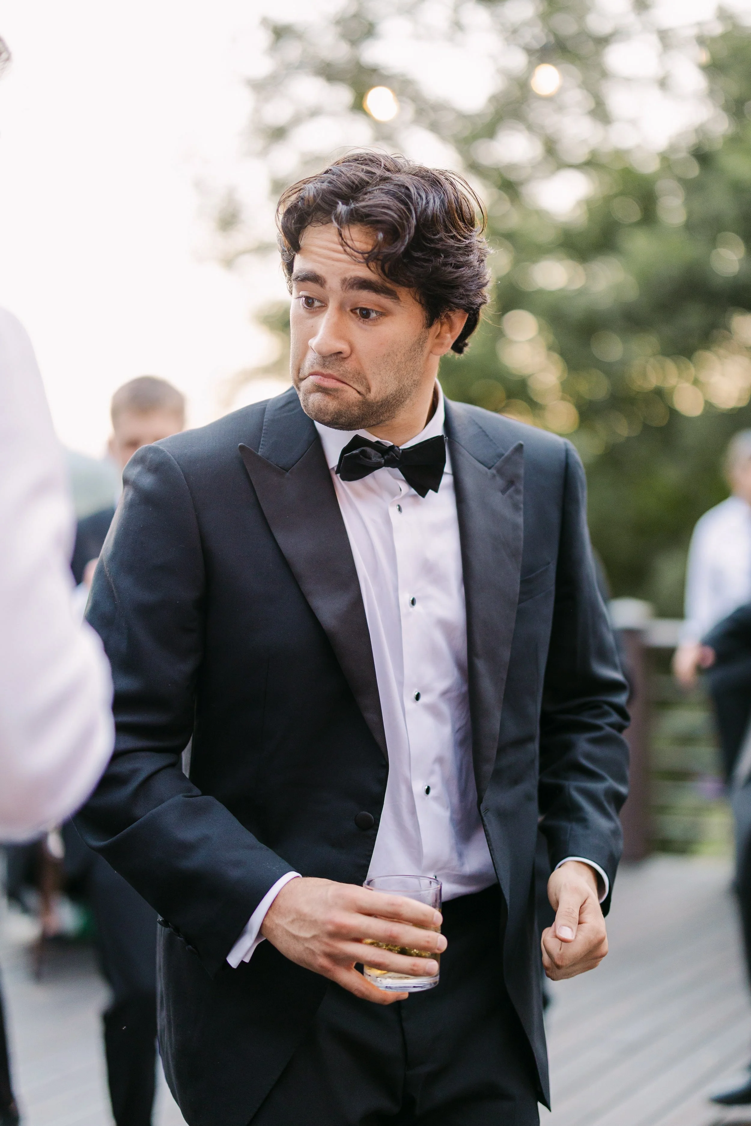 A man in a tuxedo with a bow tie holding a glass of whiskey, making a sad or unimpressed facial expression at an outdoor event.