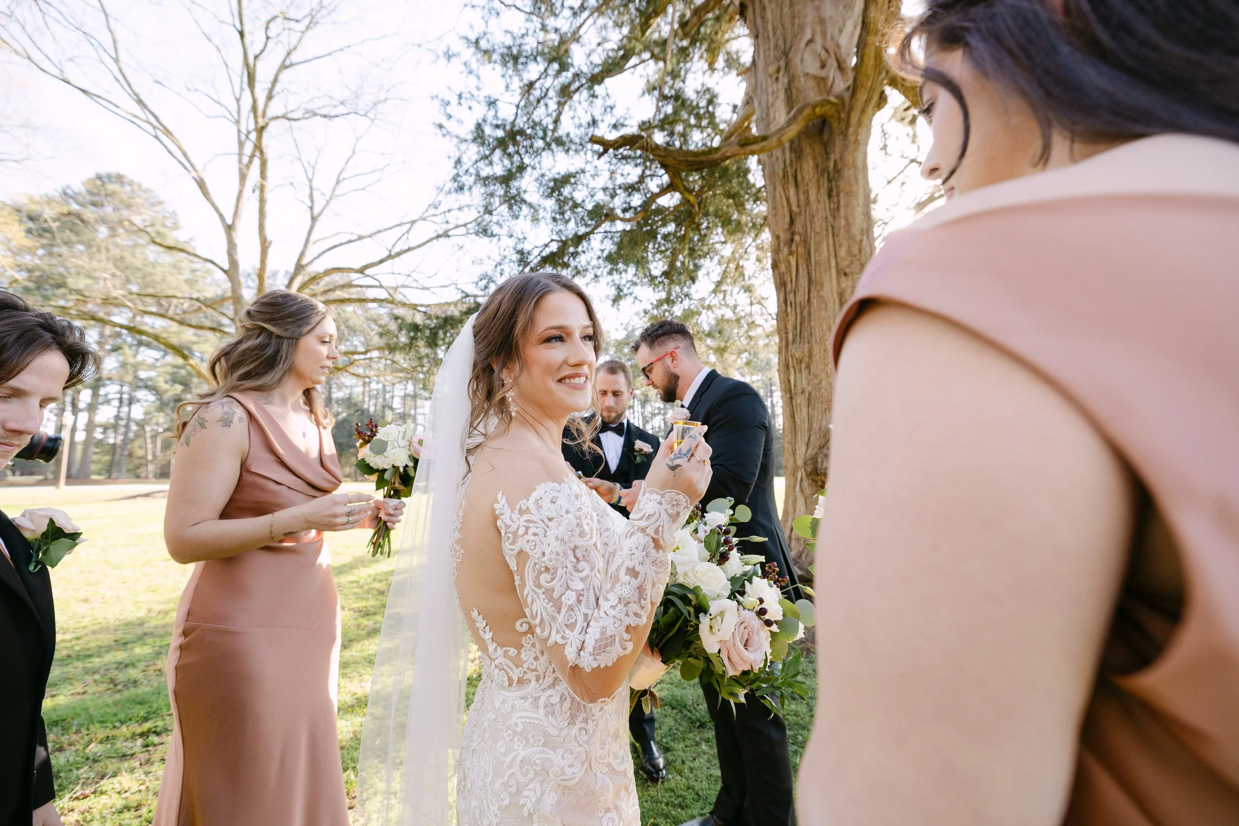 Bride holding a bouquet and smiling at her wedding ceremony outdoors, surrounded by bridesmaids and groomsmen underneath a large tree.