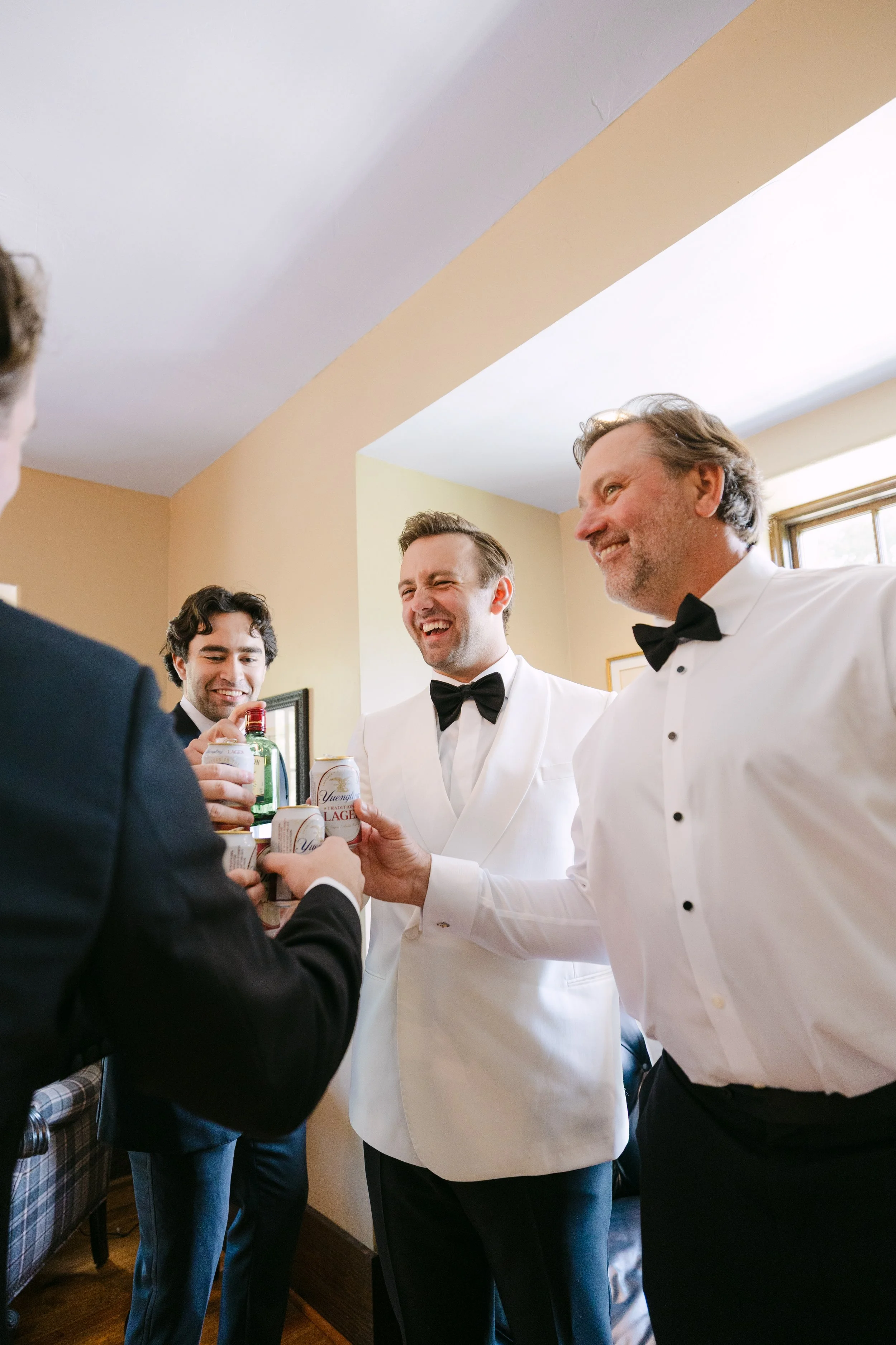 Four men in formal attire socializing indoors, some holding drinks and laughing.