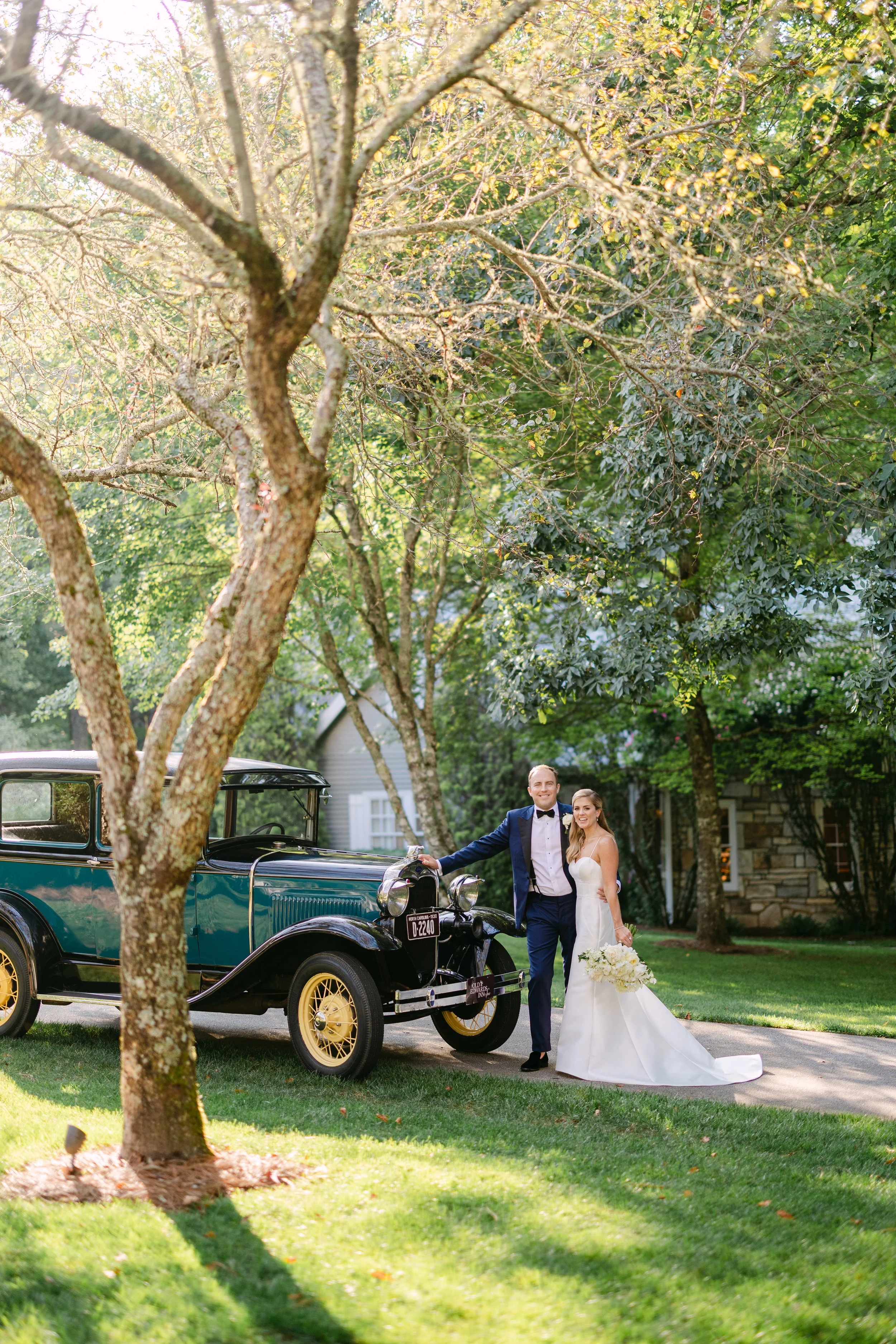 A bride in a white wedding dress holding a bouquet of white flowers and a groom in a dark suit with a bowtie standing next to a vintage blue and black car, outdoors on a grassy lawn with trees and a house in the background.
