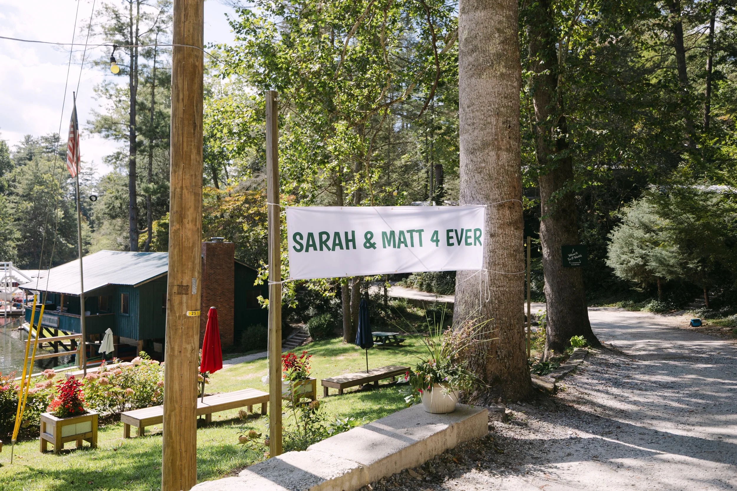 A white outdoor banner hanging between two trees reads, 'SARAH & MATT 4 EVER' near a gravel pathway, with trees, benches, and a small building in the background.