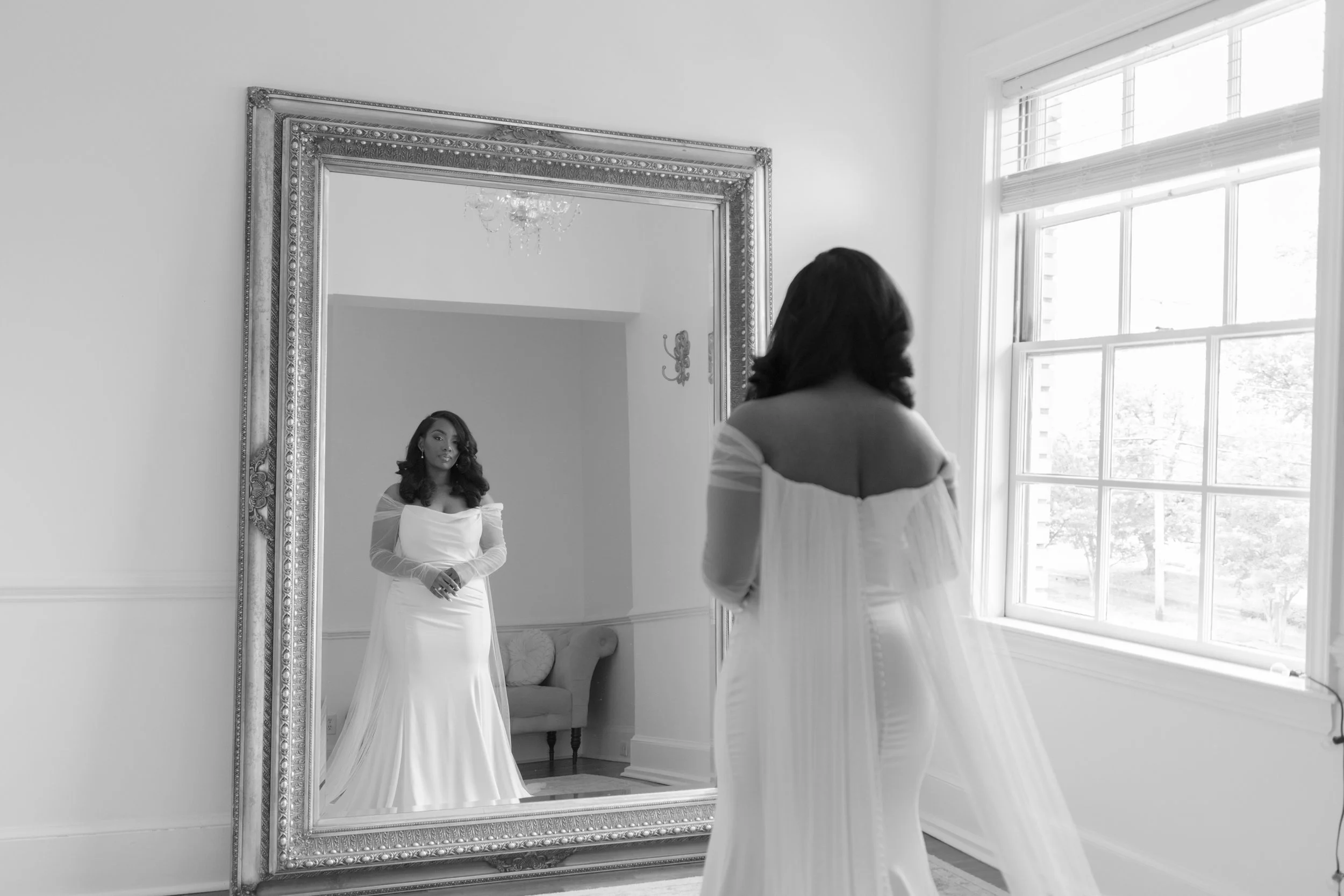 A woman in a wedding dress looking at herself in a large ornate mirror, with a window to her right letting in natural light.