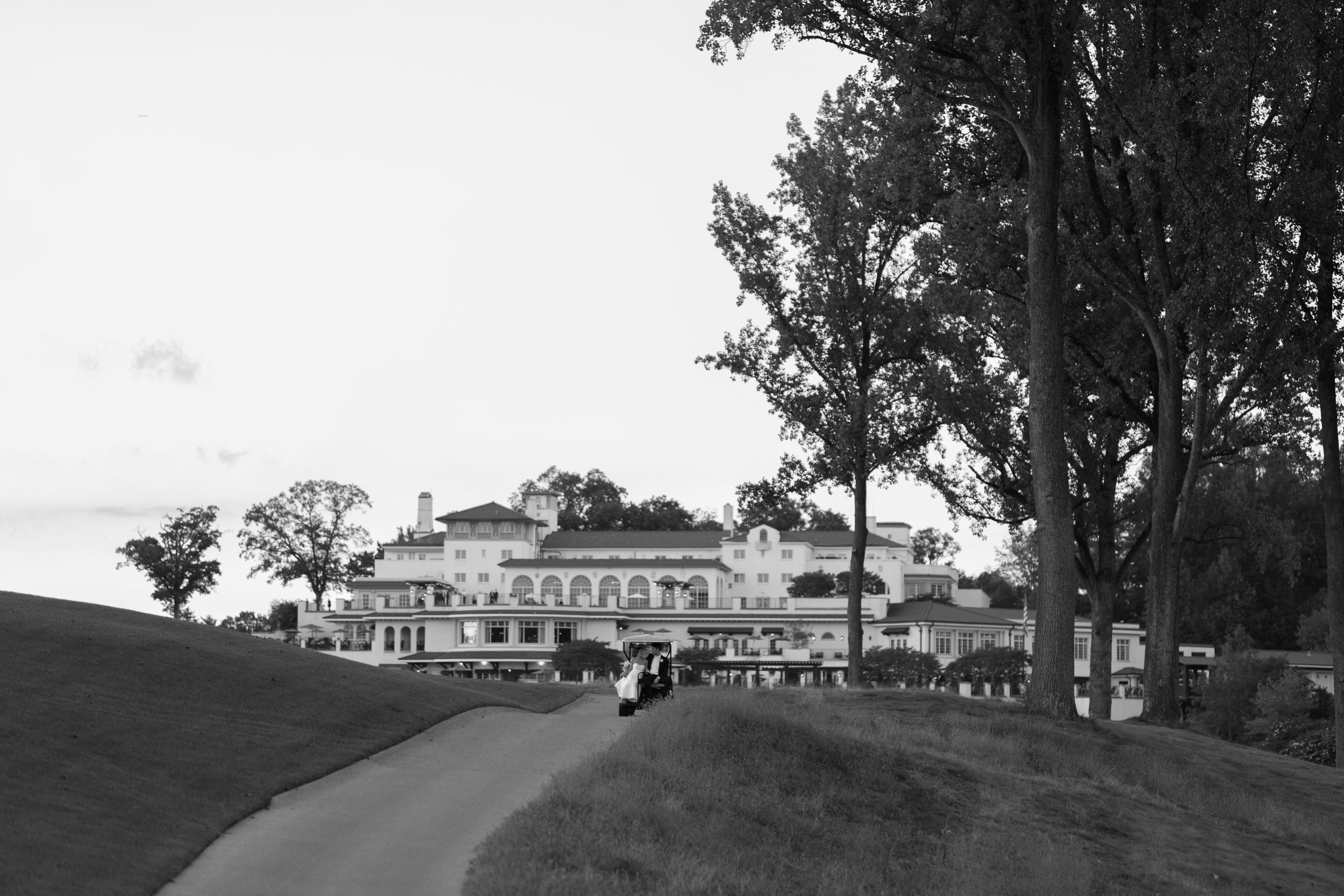 A golf cart is on a paved path leading toward a large, multi-story mansion with numerous windows, balconies, and architectural details, overlooking a golf course with trees on both sides.