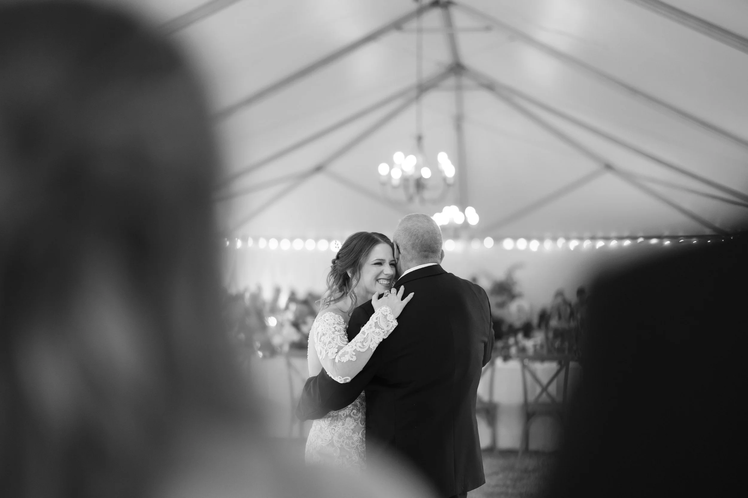 Black and white photo of a bride and groom dancing, smiling and embracing, inside a decorated wedding tent.
